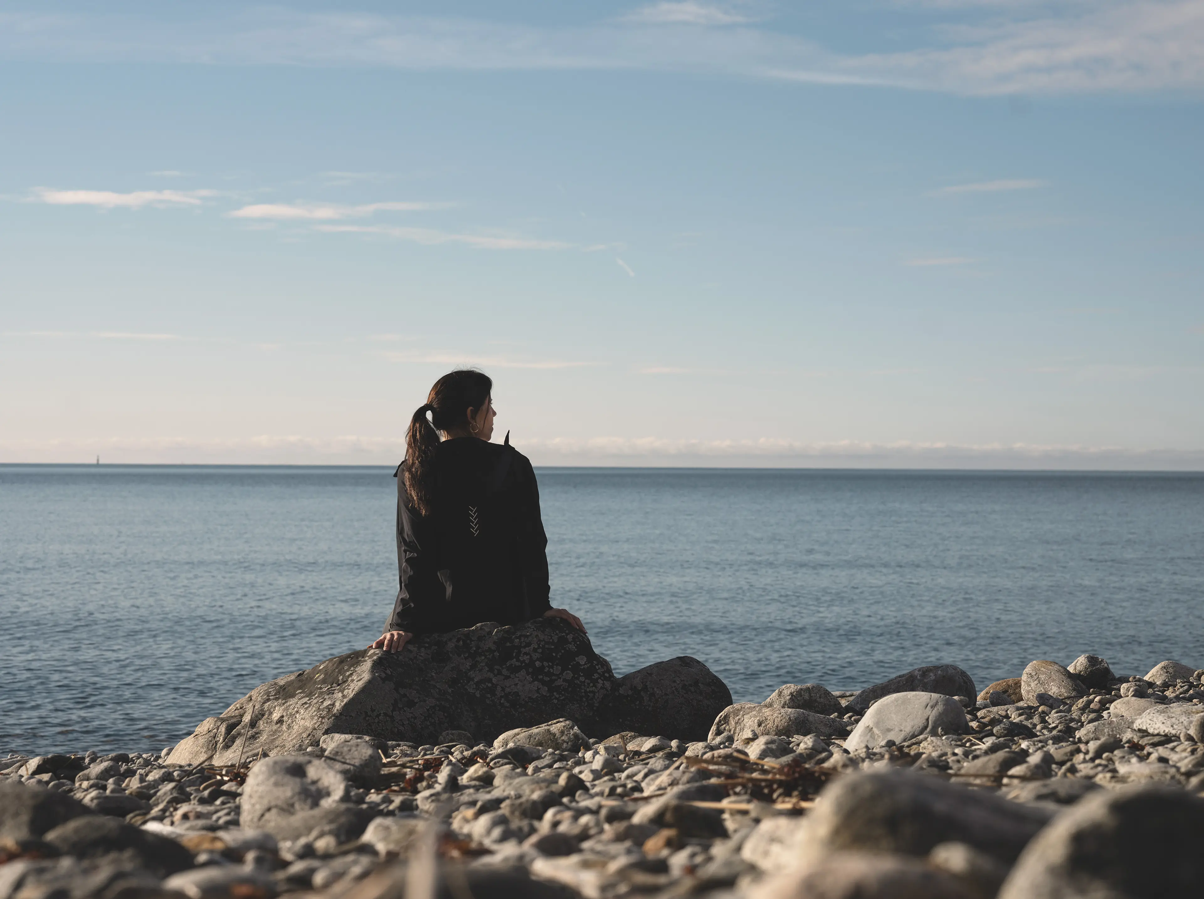 A person sitting on a rocky beach, facing the calm sea under a clear blue sky.