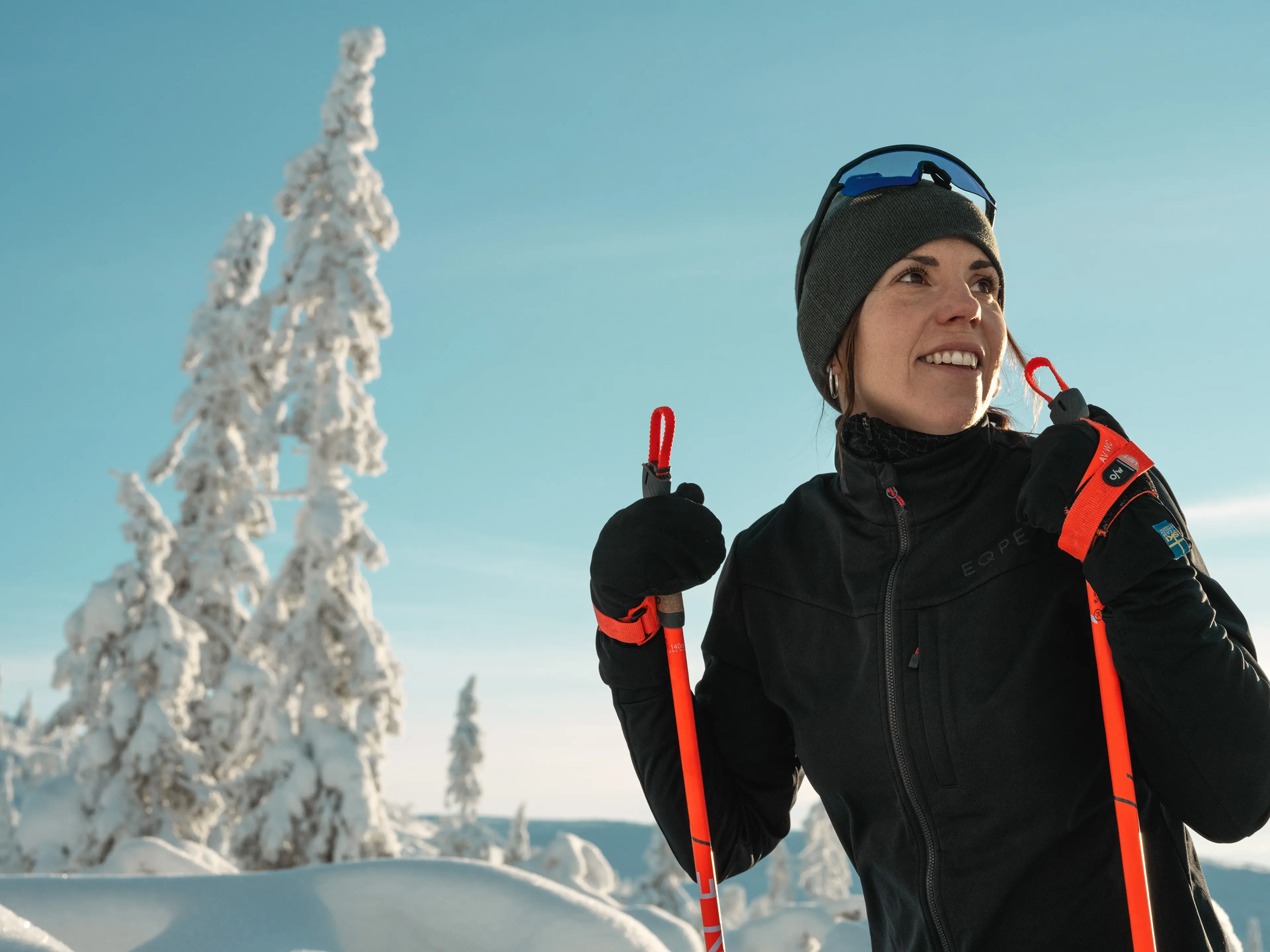 Person in winter gear holding ski poles, smiling in a snowy landscape with frosted trees and clear blue sky.