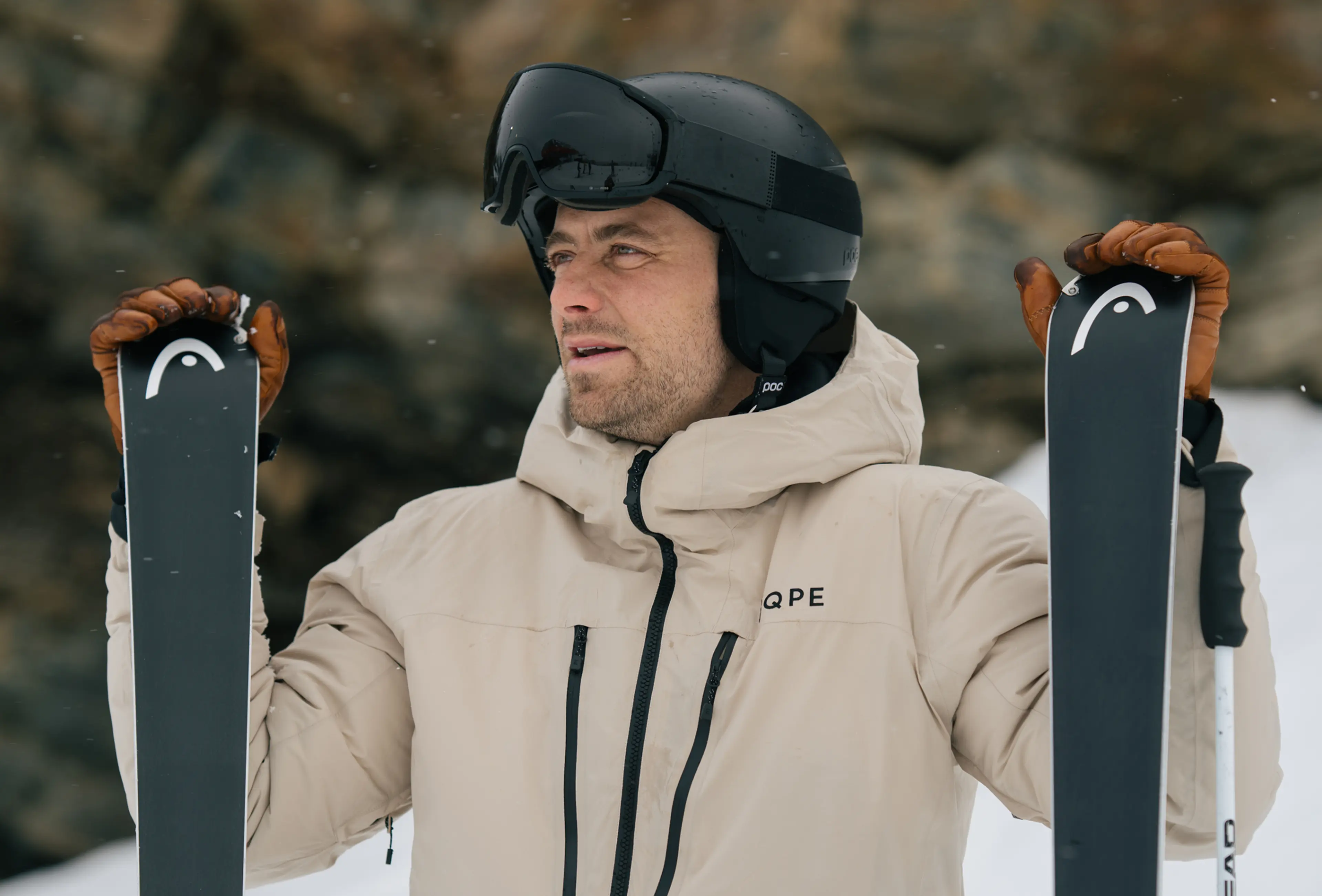 A skier in a beige jacket and black helmet holds skis, standing in a snowy landscape.