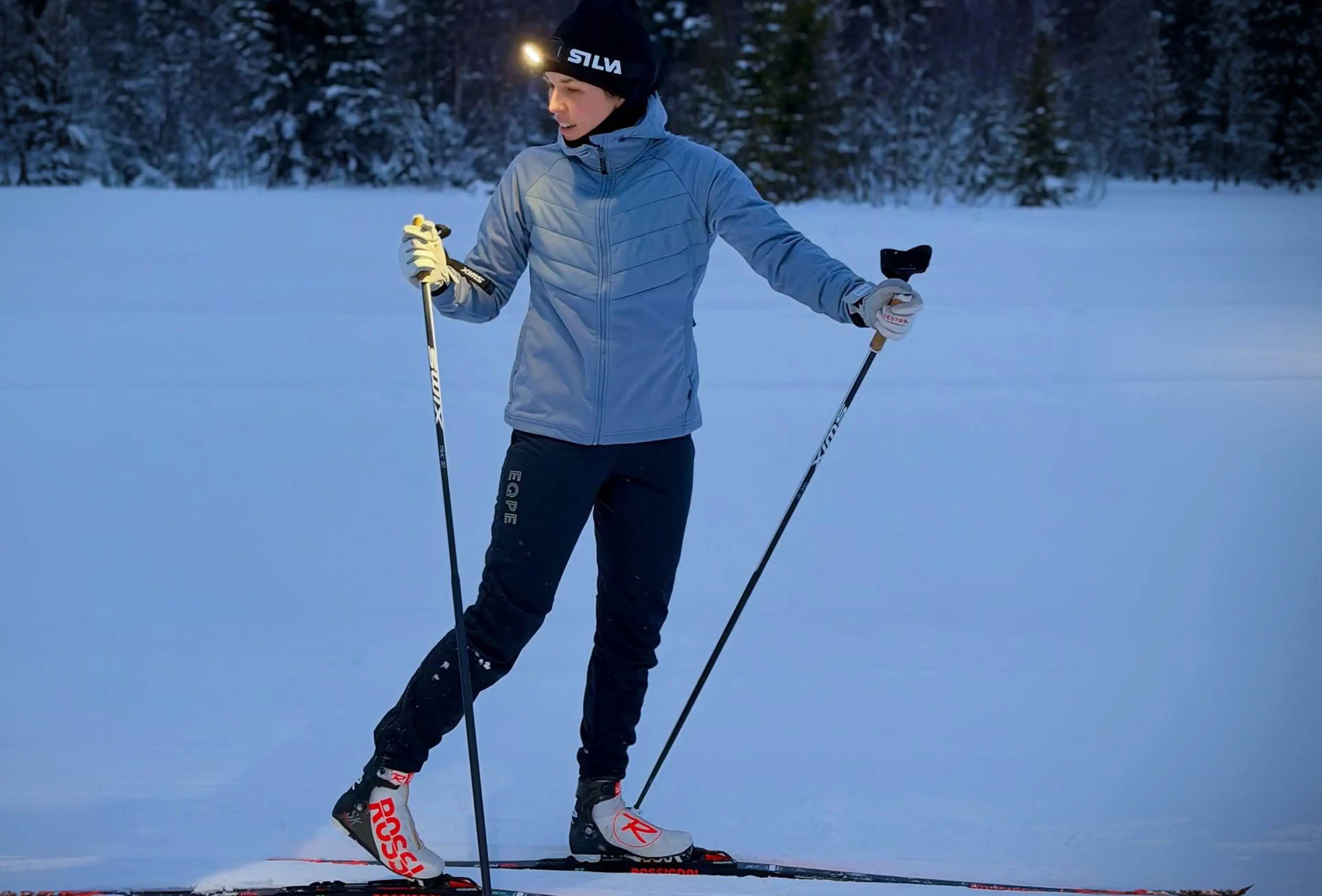 Person cross-country skiing on a snowy trail, wearing a headlamp and winter gear, surrounded by a snowy forest landscape.