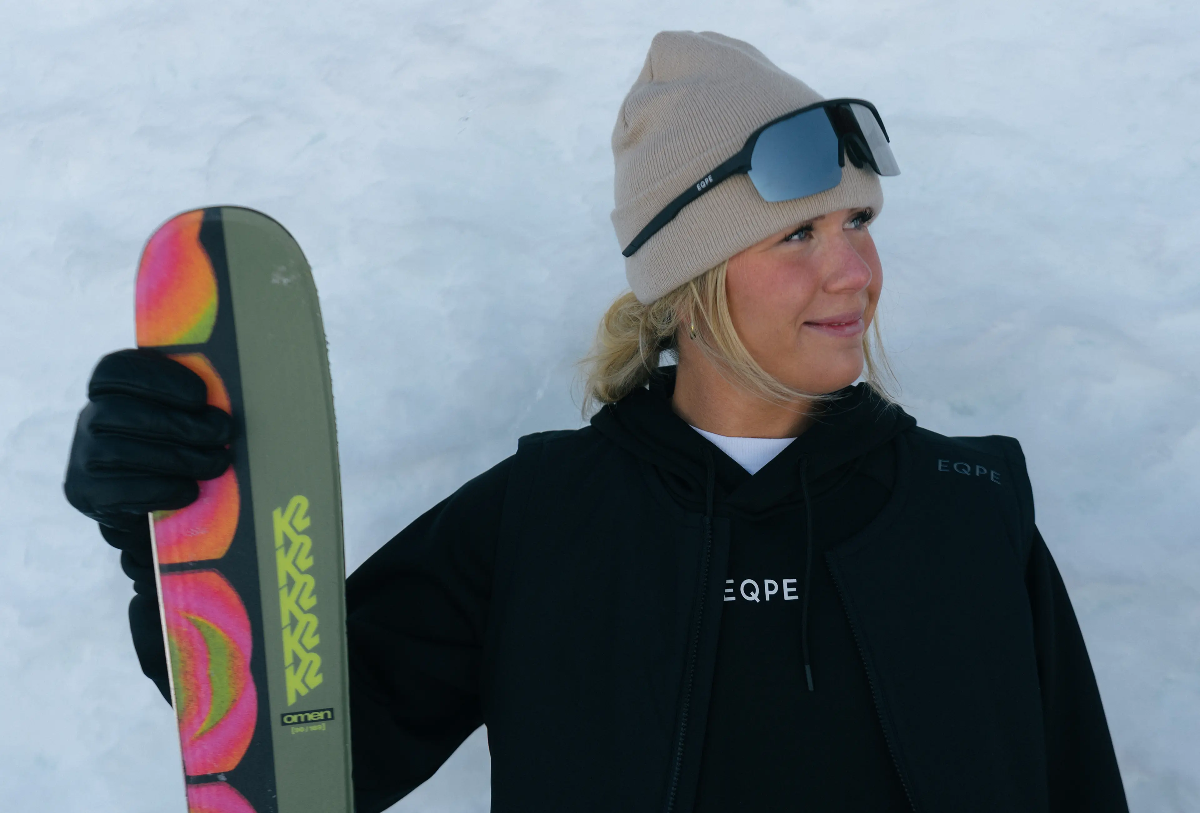 A person wearing a beige beanie and sunglasses holds a colorful ski against a snowy background, smiling and looking to the side.