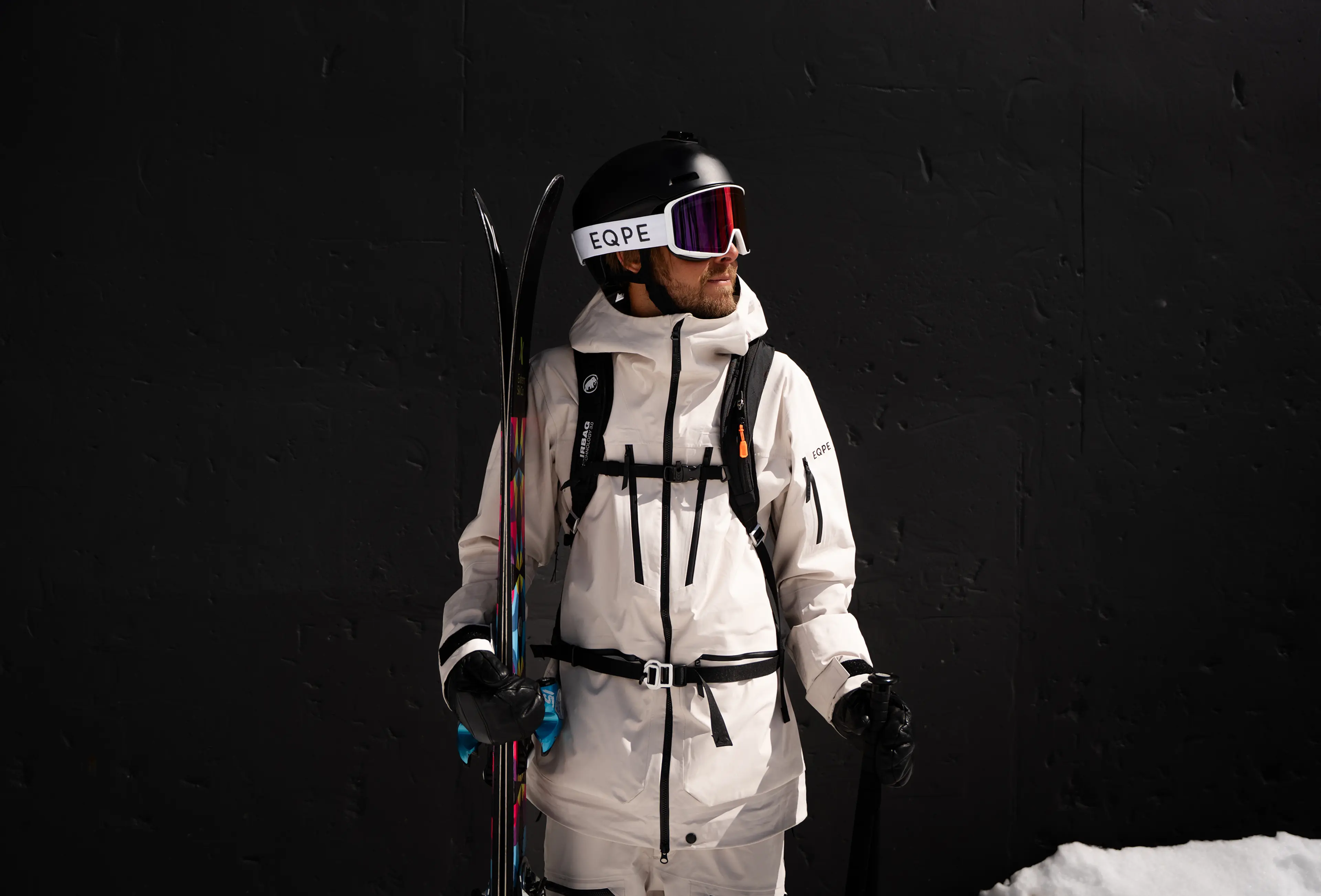 A skier in a white ski outfit holds skis and poles against a dark background, wearing a helmet and goggles.