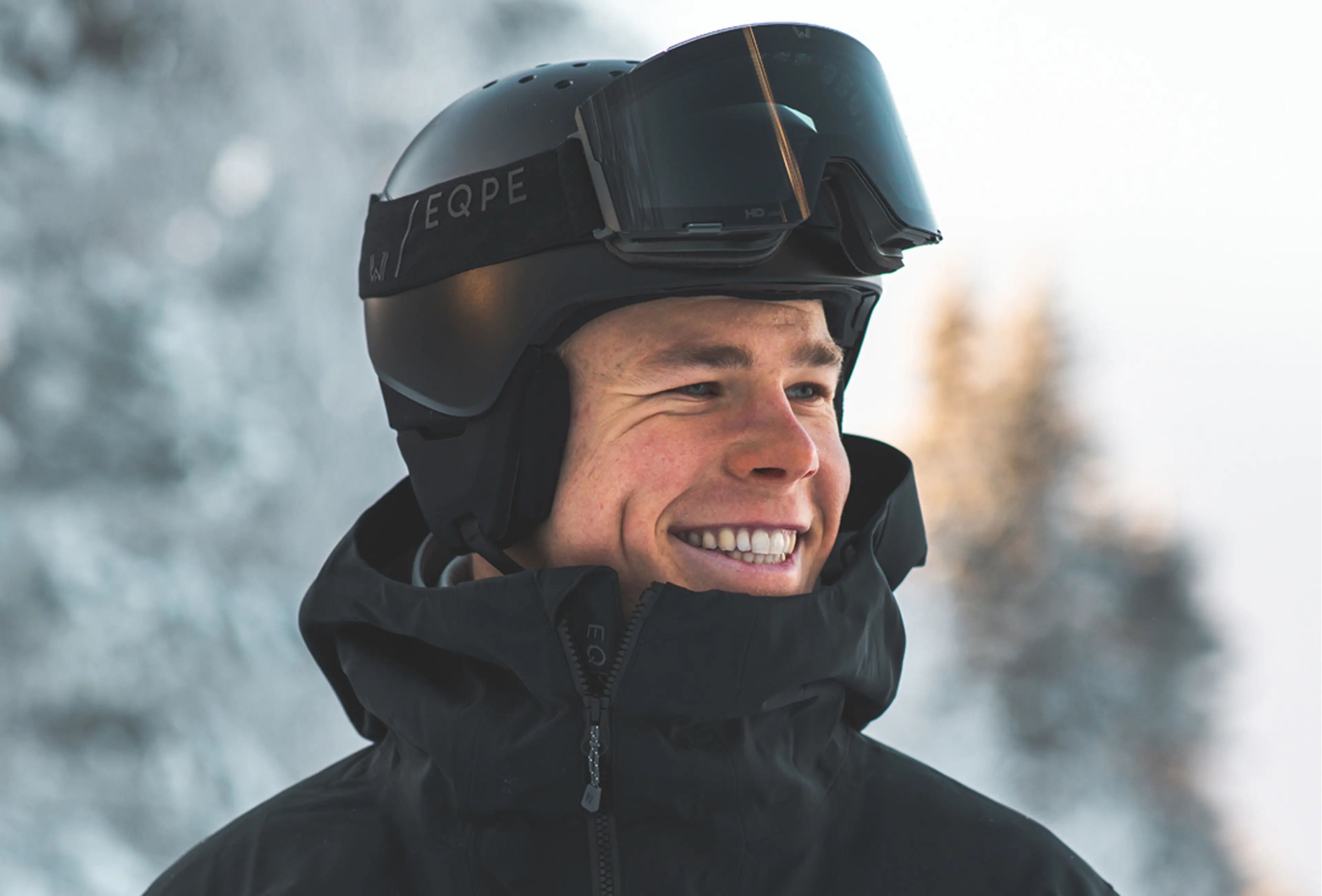 Smiling person wearing a black helmet and goggles, standing outdoors with a snowy background.