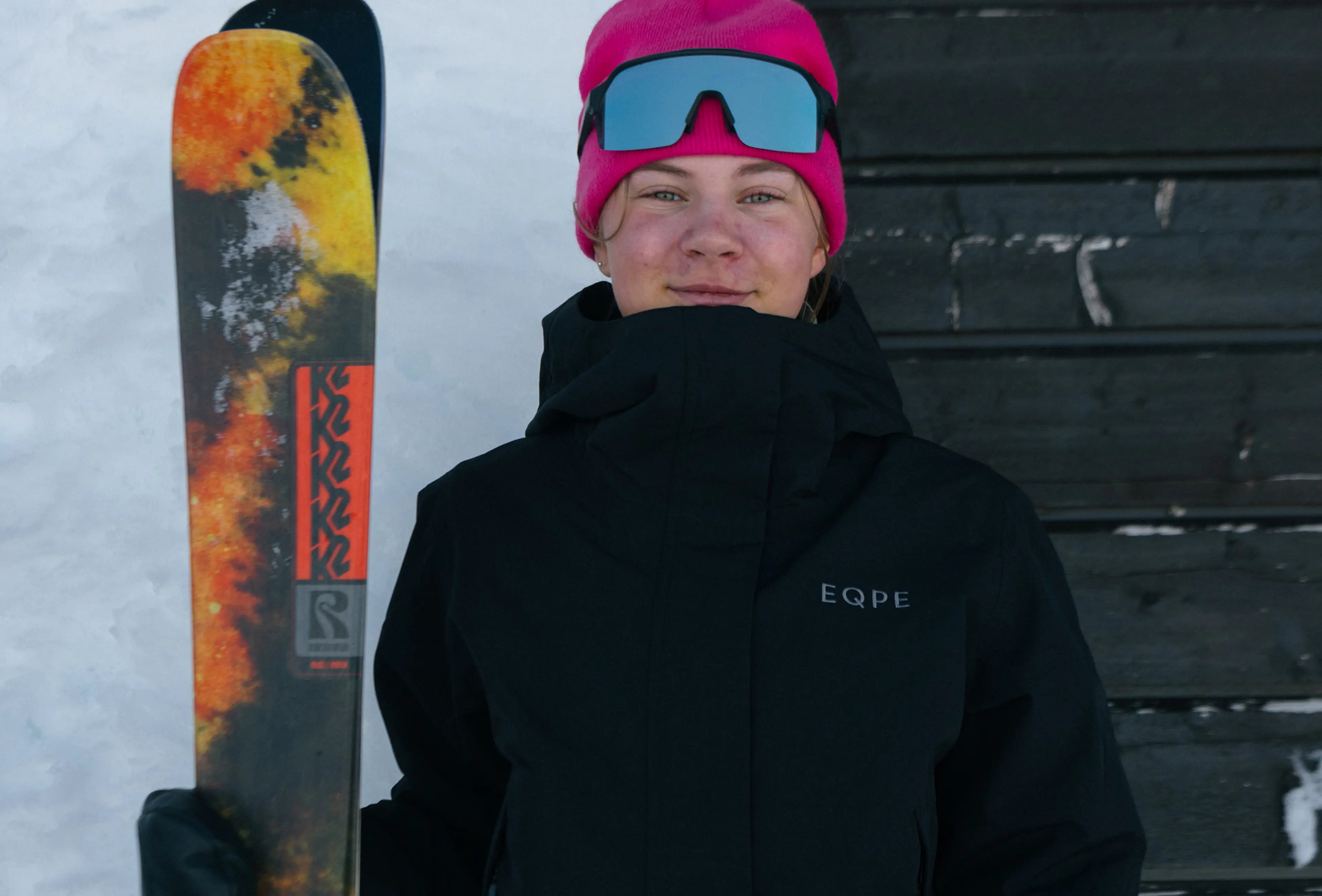 Person in a black jacket and pink beanie holds a colorful ski, standing against a snow-covered background.