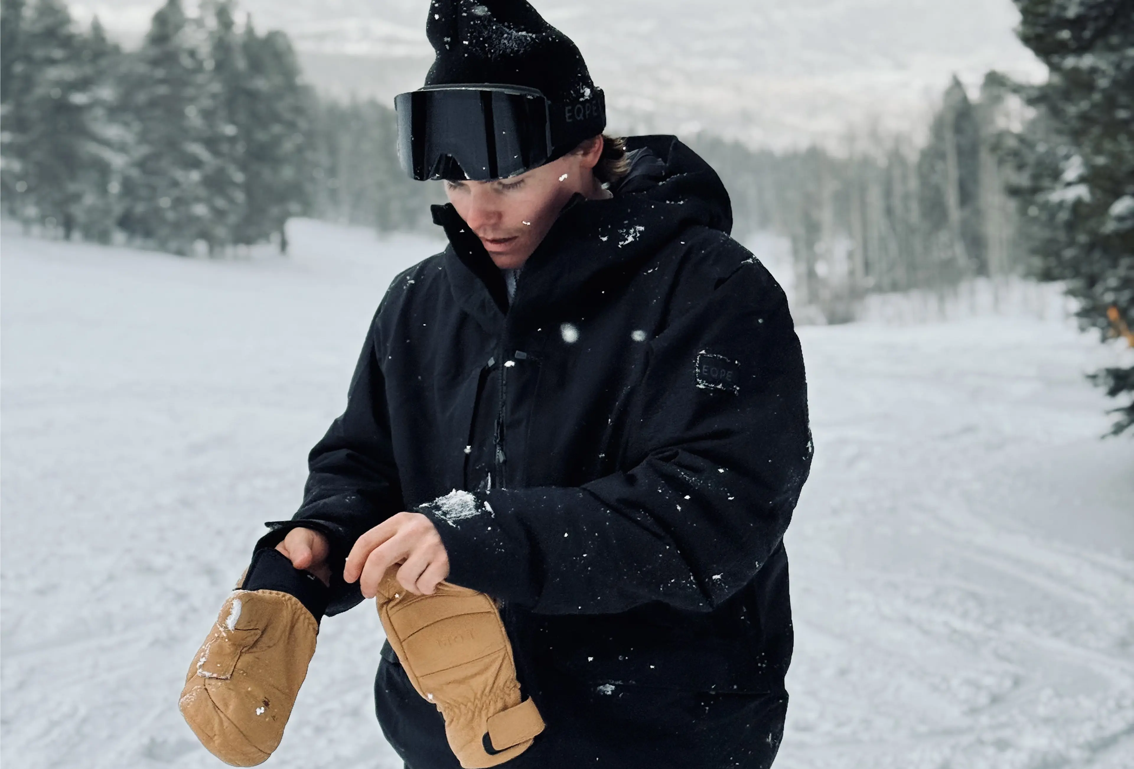 Person in black winter gear and goggles adjusting tan mittens in a snowy, tree-lined landscape.