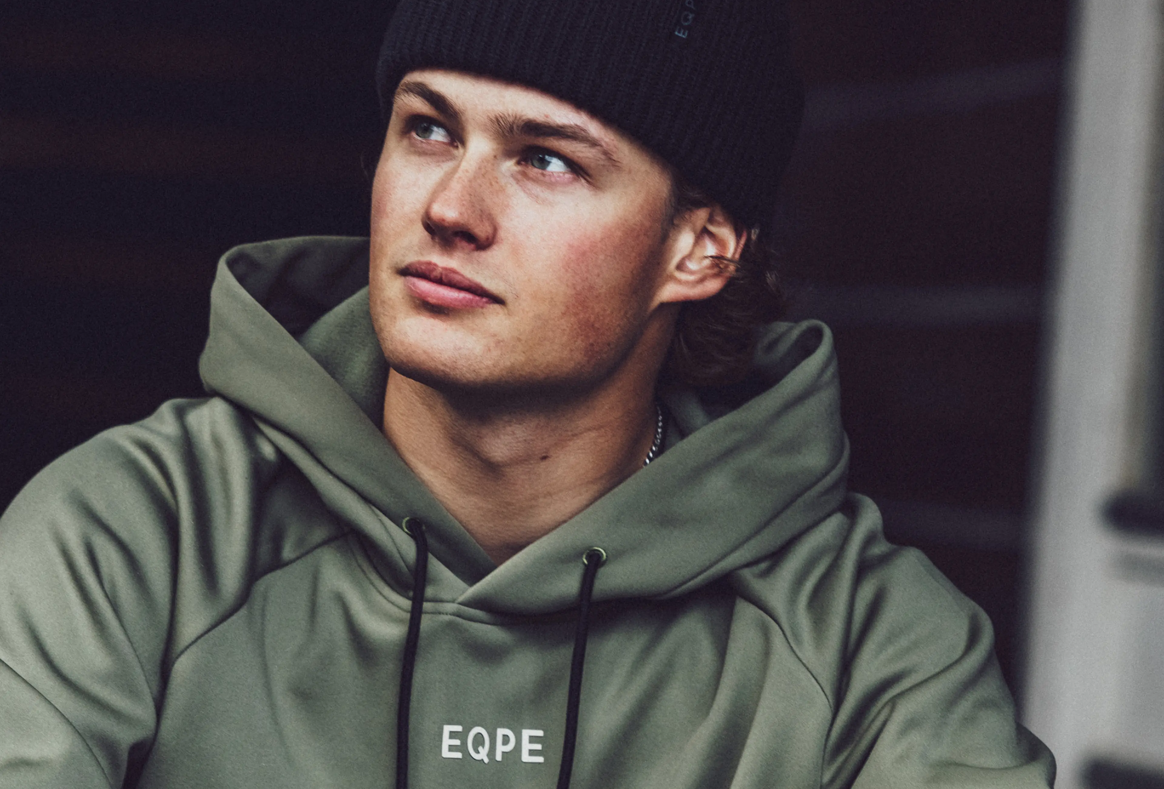 Person wearing a green hoodie and black beanie, gazing thoughtfully to the side against a dark background.