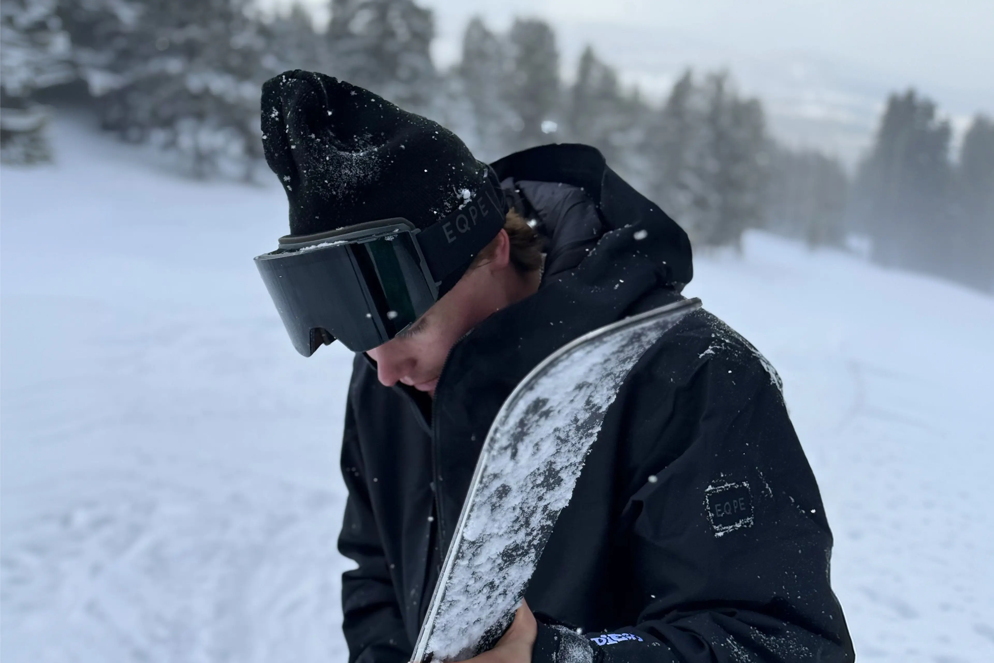 Person in black snow gear and goggles holding a snow-covered snowboard in a snowy, forested landscape.