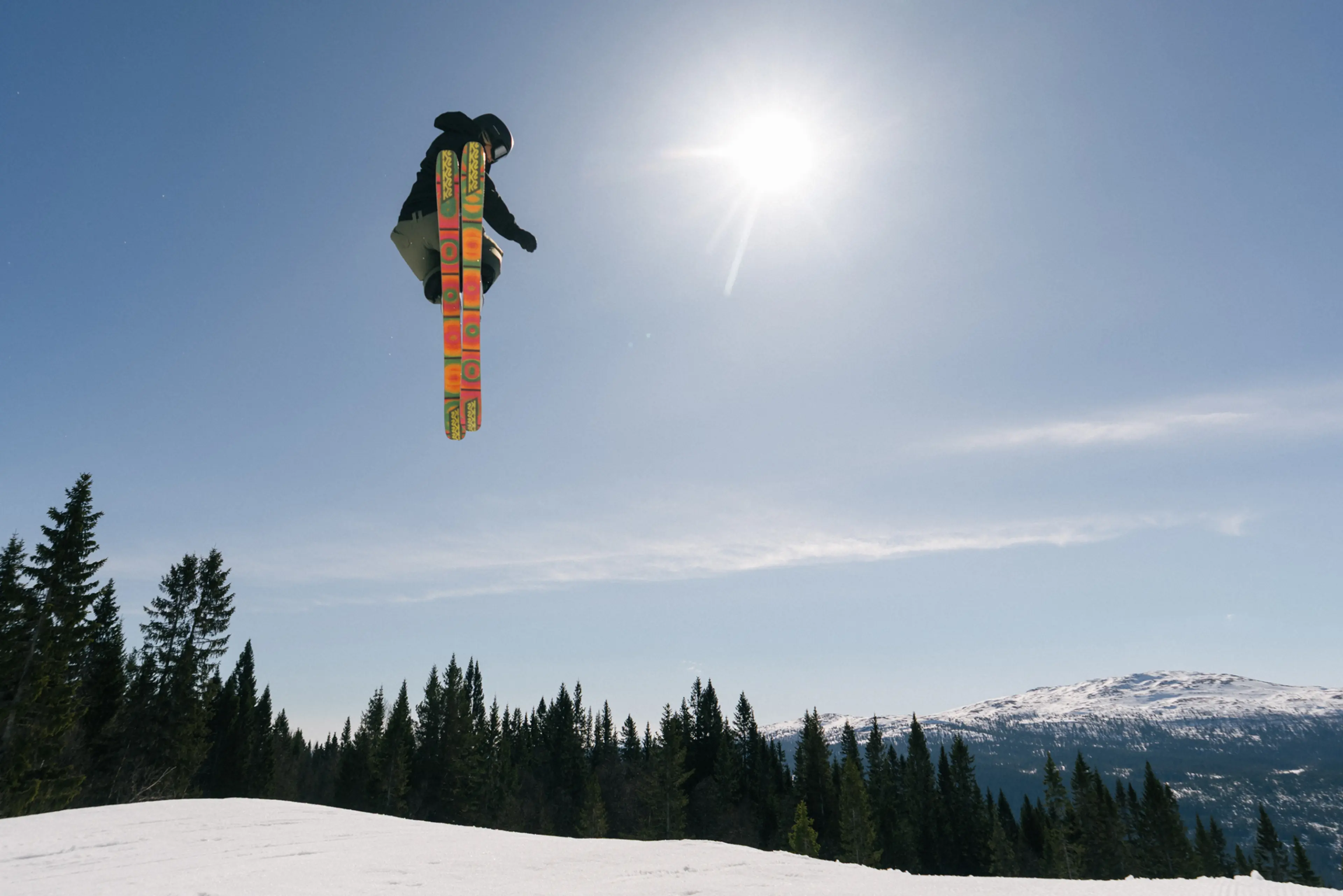 A skier in mid-air performs a jump against a clear blue sky, with snow-covered mountains and trees in the background.
