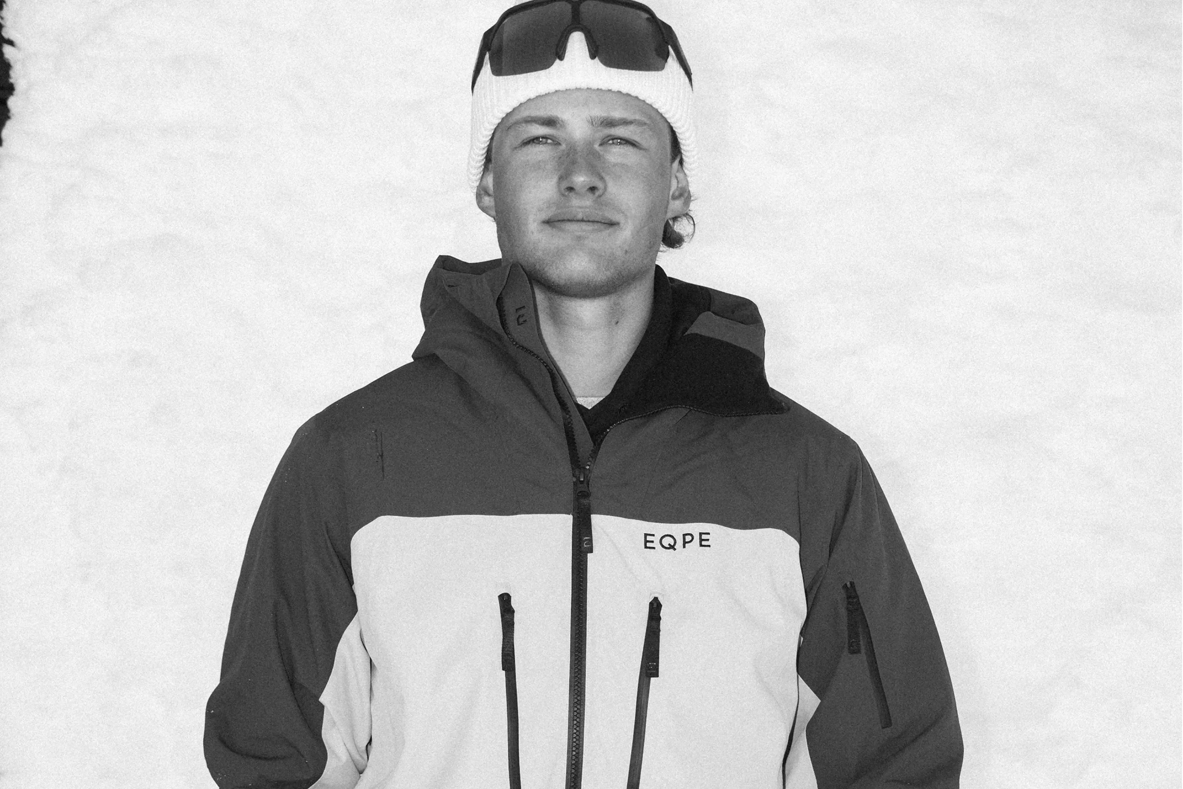 Person in a winter jacket and beanie with sunglasses on head, standing against a snowy background.