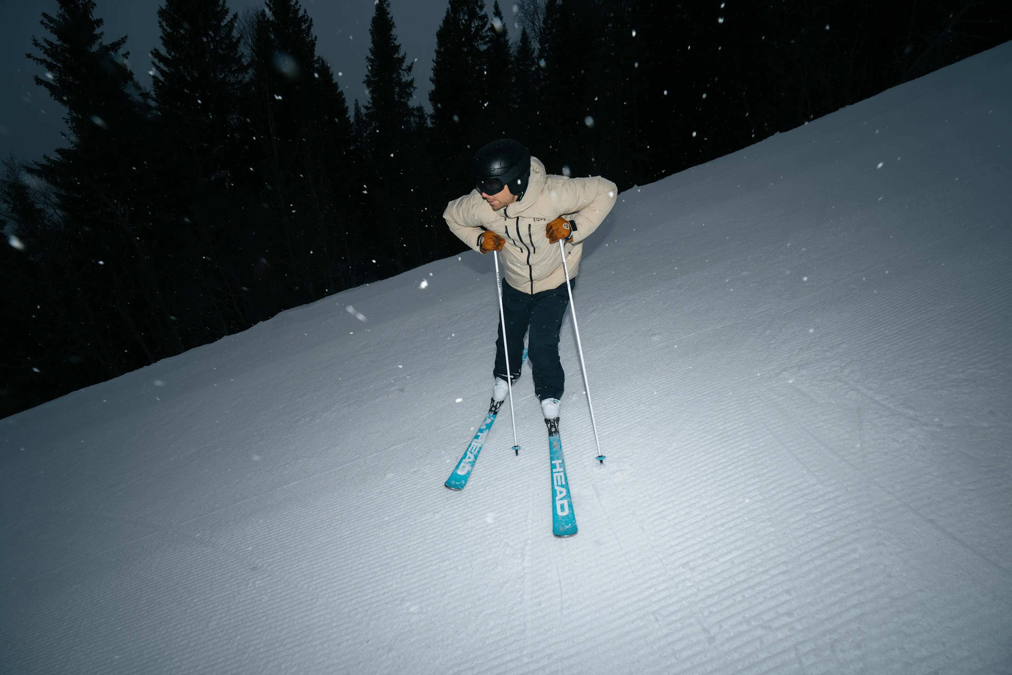 A skier in a beige jacket and helmet navigates down a snowy slope, with snowflakes falling and pine trees in the background.