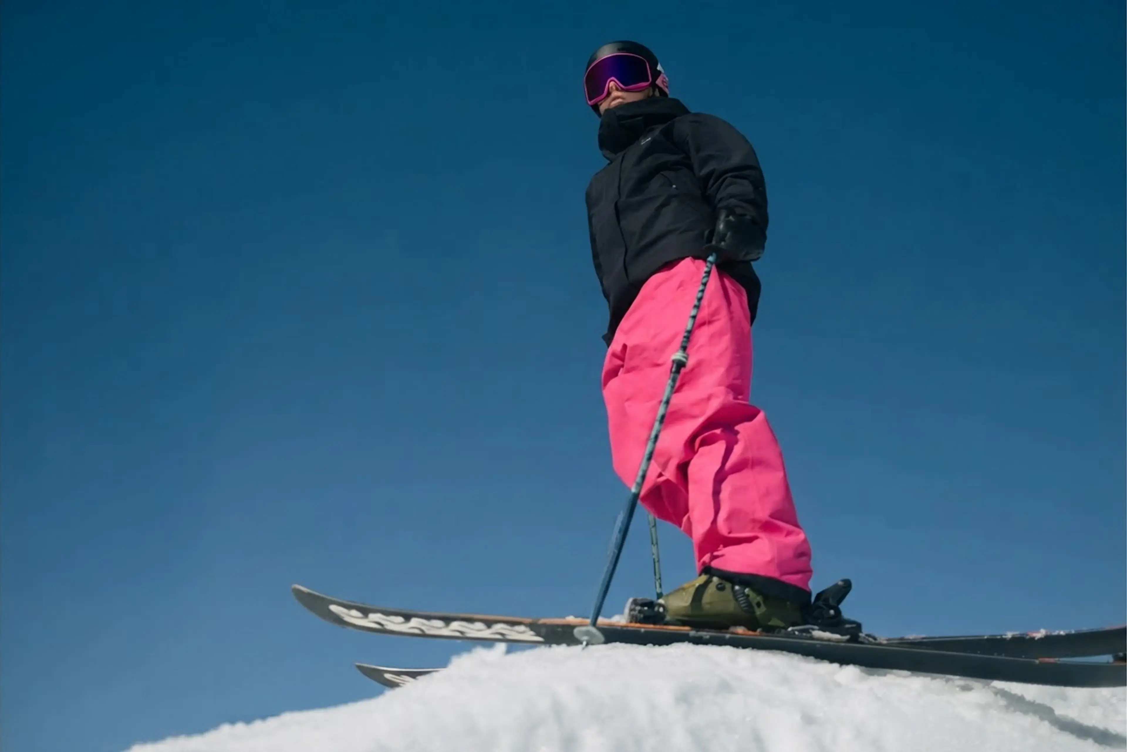 Skier in bright pink pants and black jacket stands atop a snowy slope, holding ski poles, with a clear blue sky in the background.