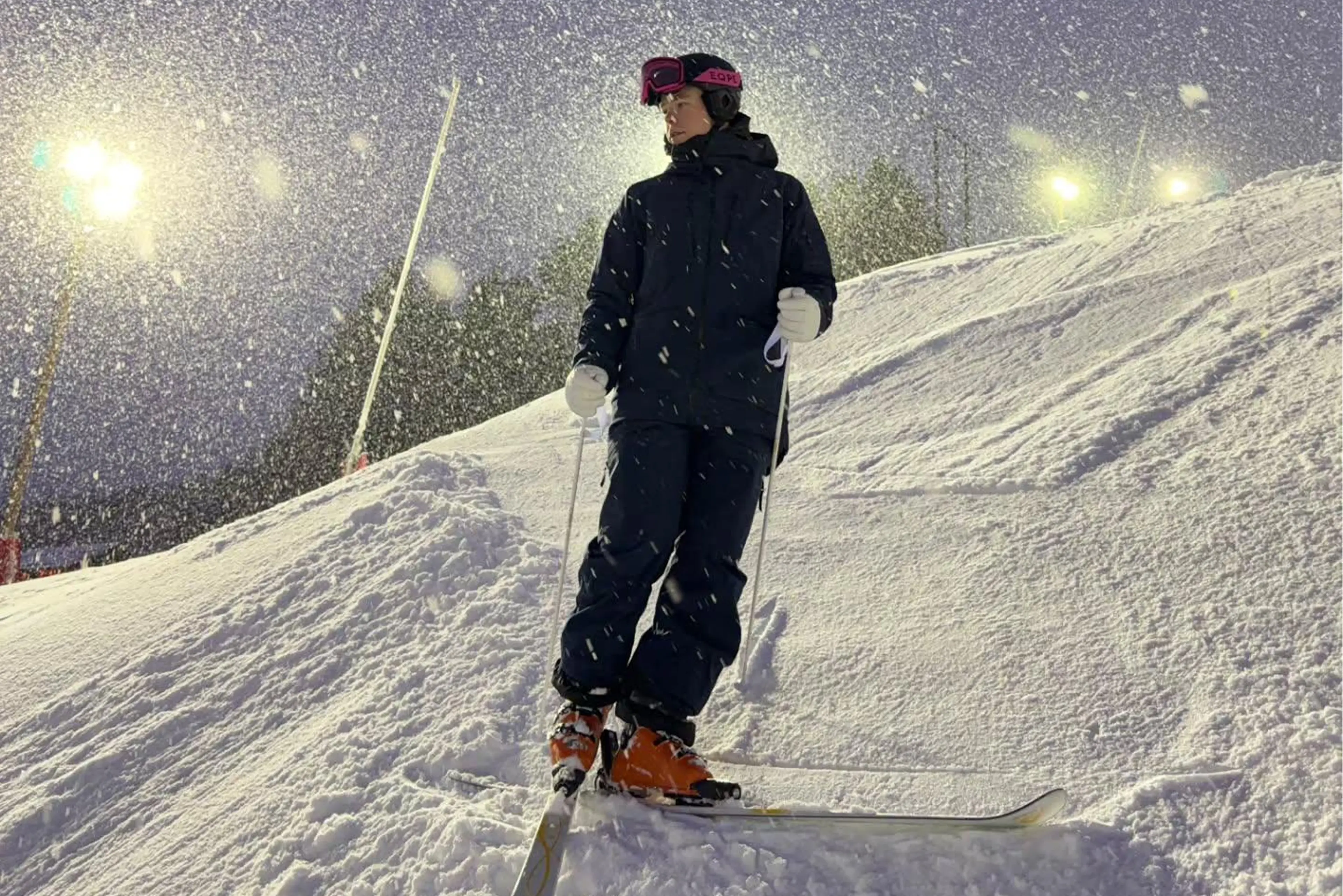 A skier in dark winter gear stands on a snowy slope at night, surrounded by falling snow and illuminated by bright lights.