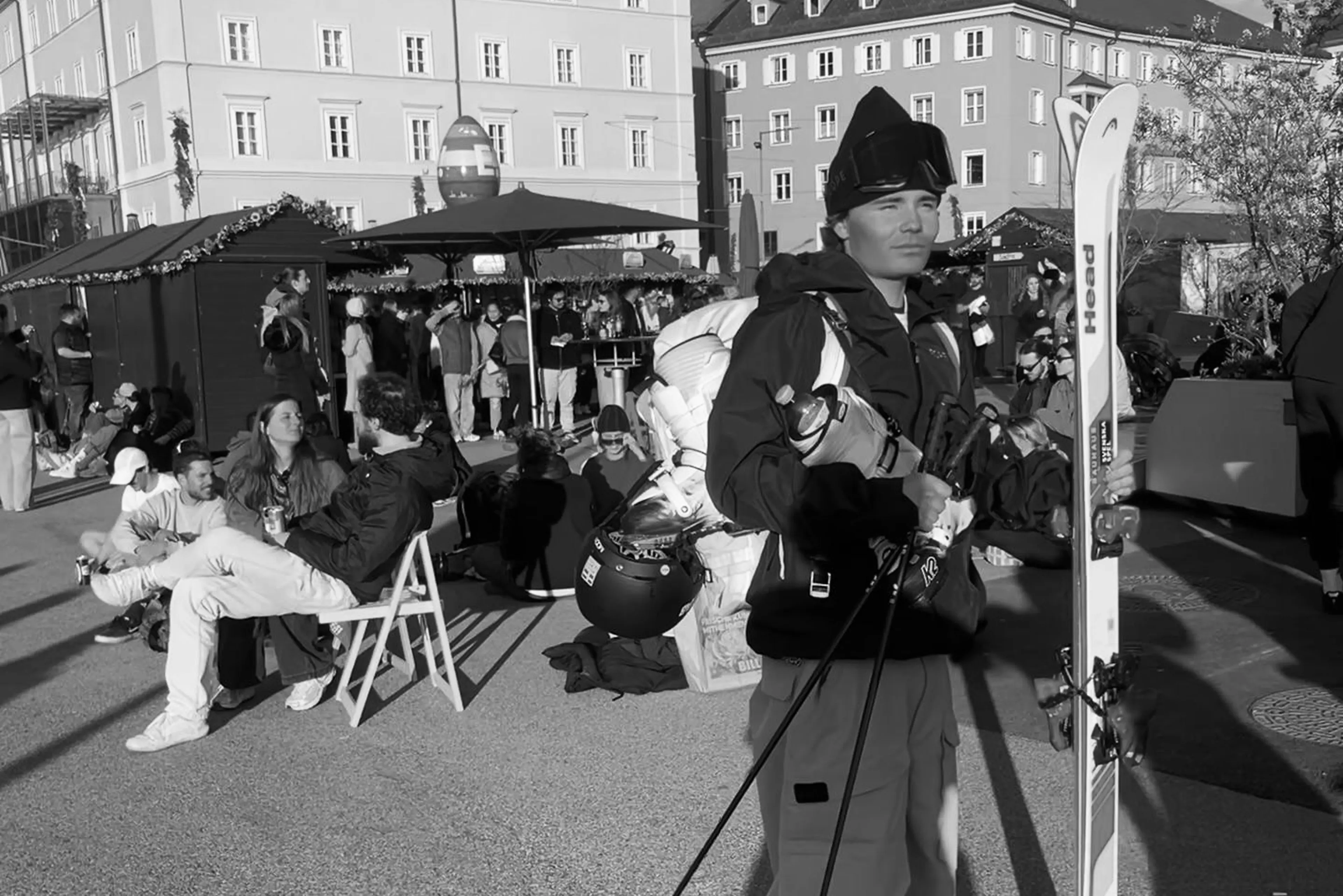 A person in ski gear stands with skis and poles near a busy outdoor cafe, surrounded by people seated and socializing.