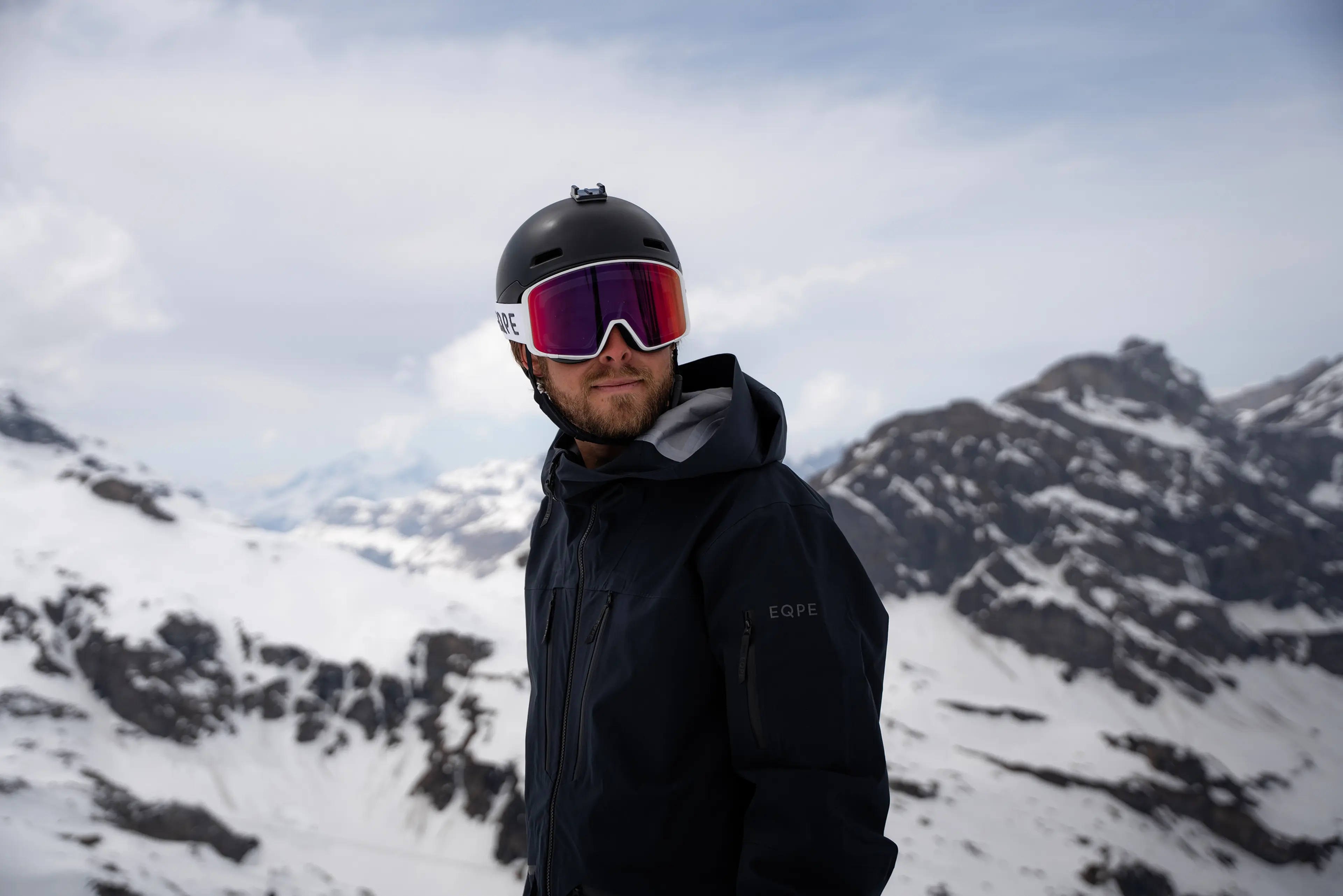 A person wearing a black helmet and goggles stands against a snowy mountain backdrop, dressed in a black jacket.
