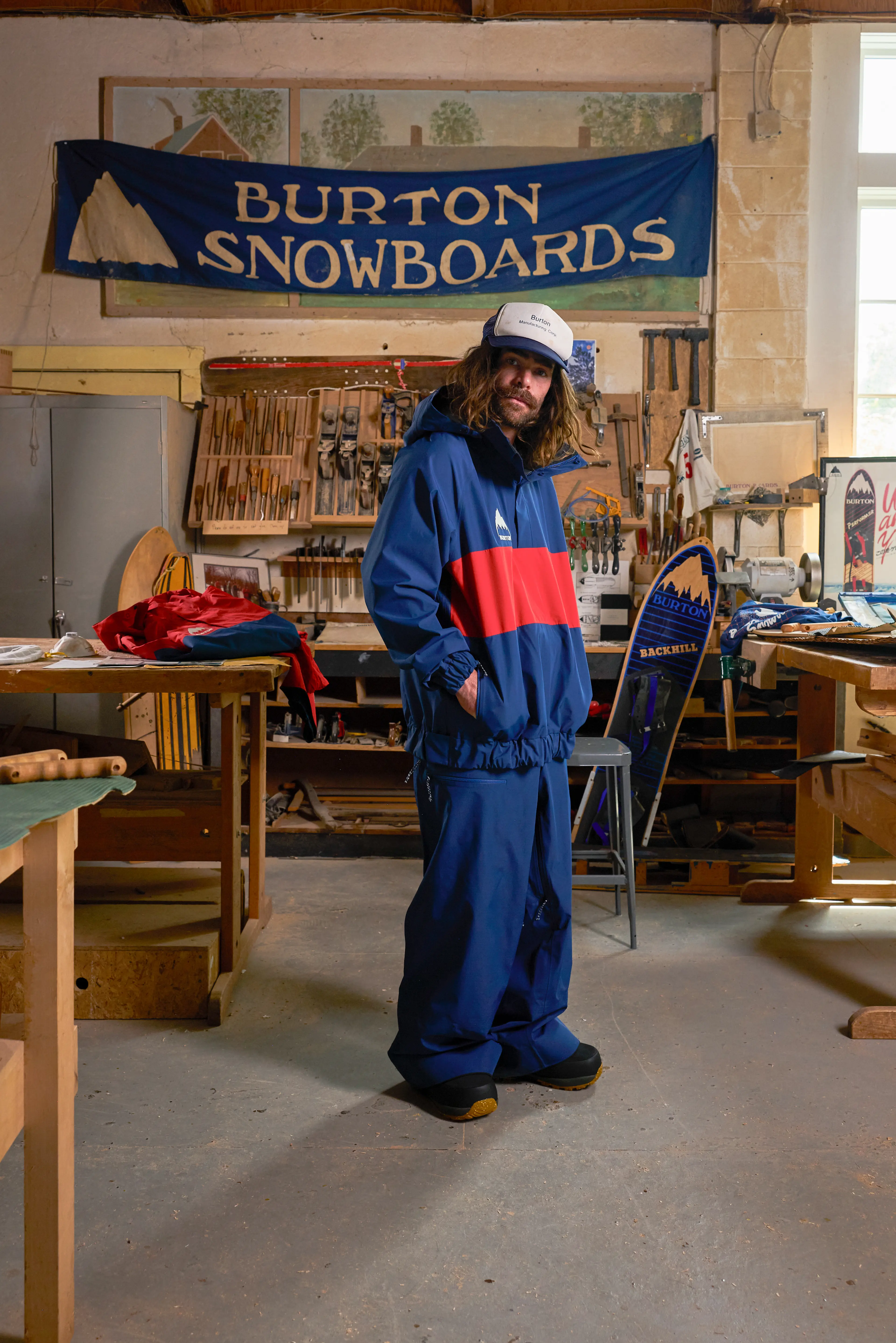 A person in a blue and red snowsuit stands in a workshop with a "Burton Snowboards" banner and woodworking tools in the background.