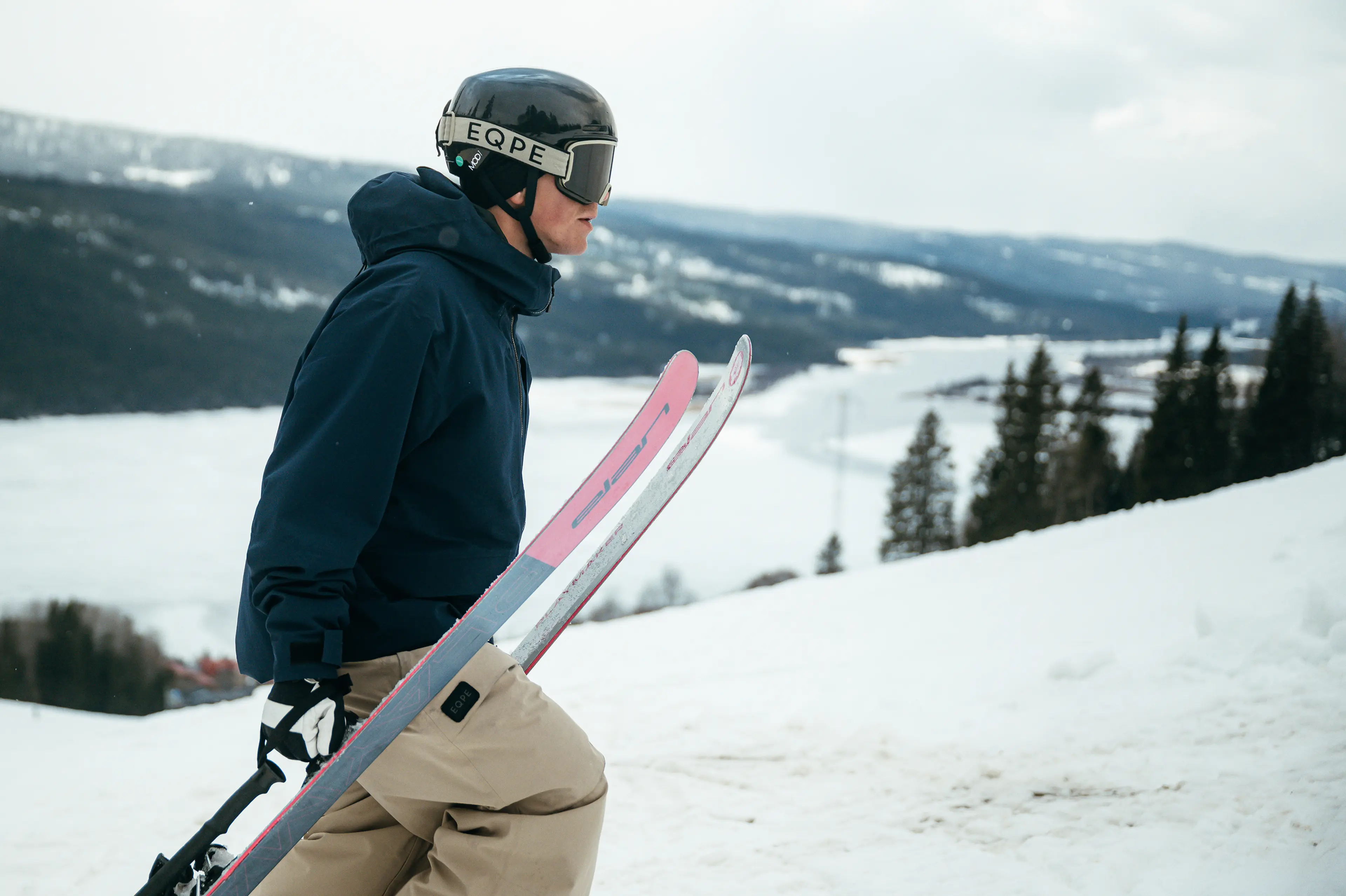 A person in ski gear holds pink skis, standing on a snowy slope with a mountainous landscape in the background.