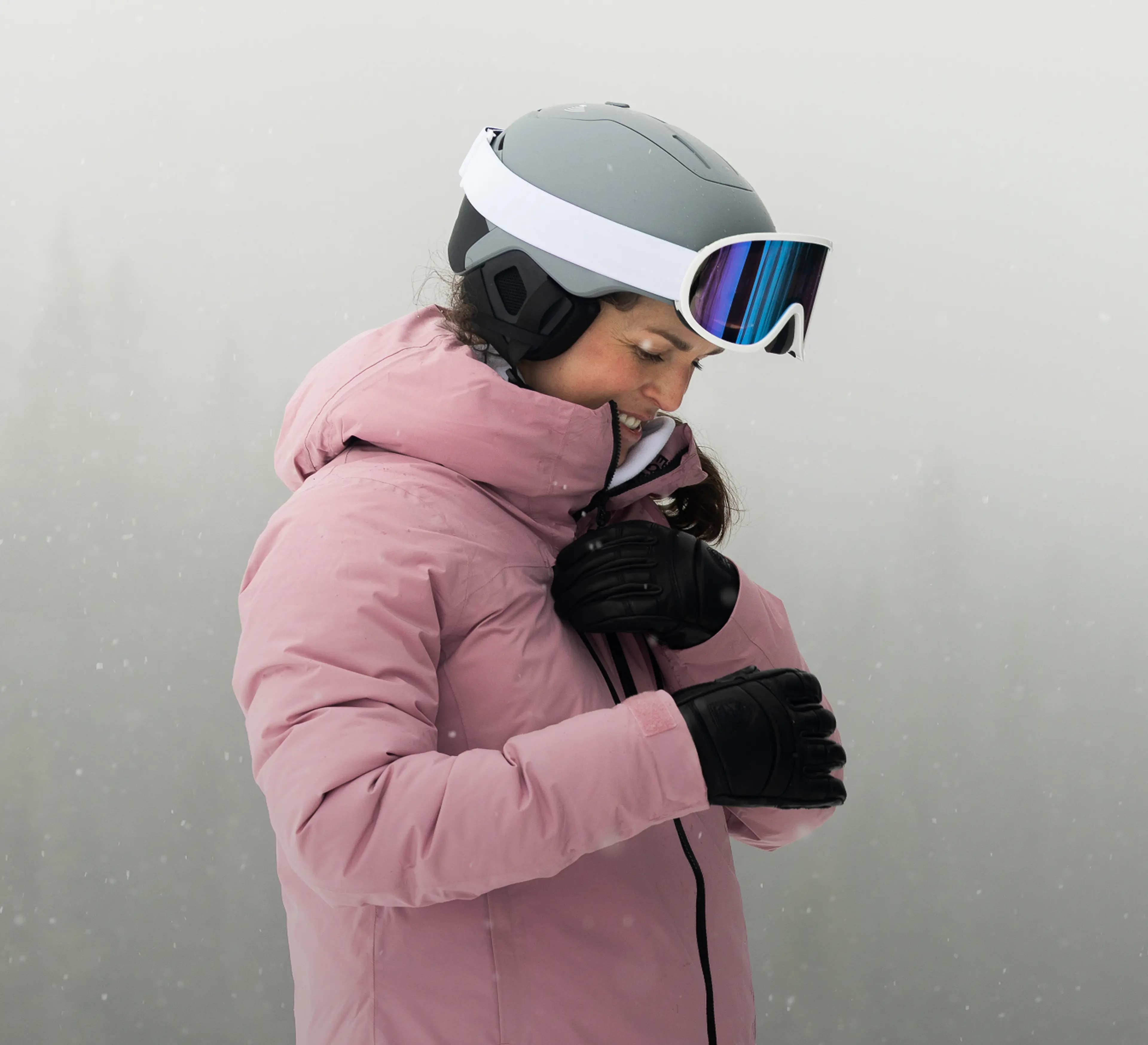 Person in a pink jacket and ski helmet adjusting their collar in a snowy, foggy setting.