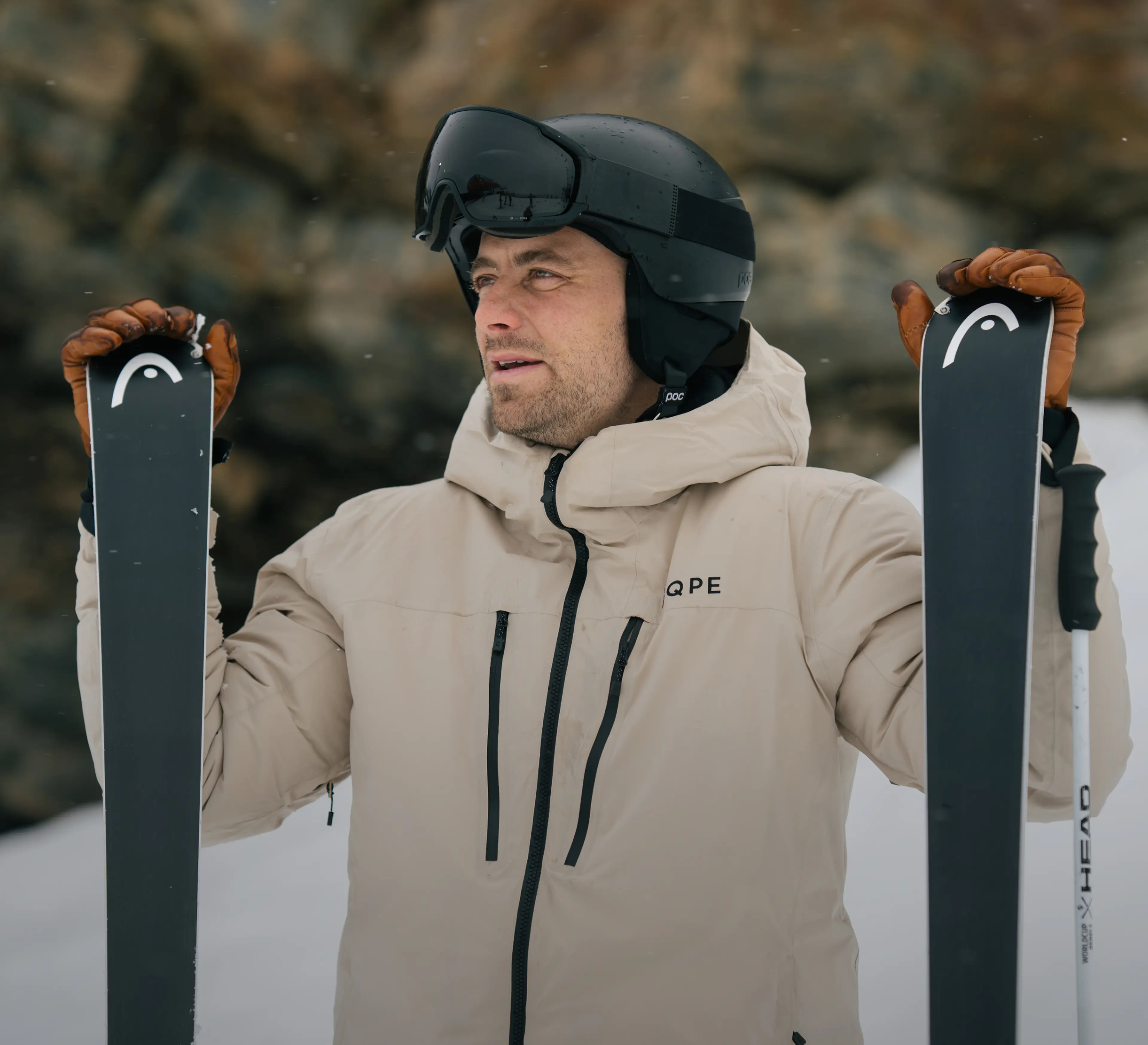 A skier in a beige jacket and black helmet holds skis, gazing into the distance in a snow-covered, rocky landscape.