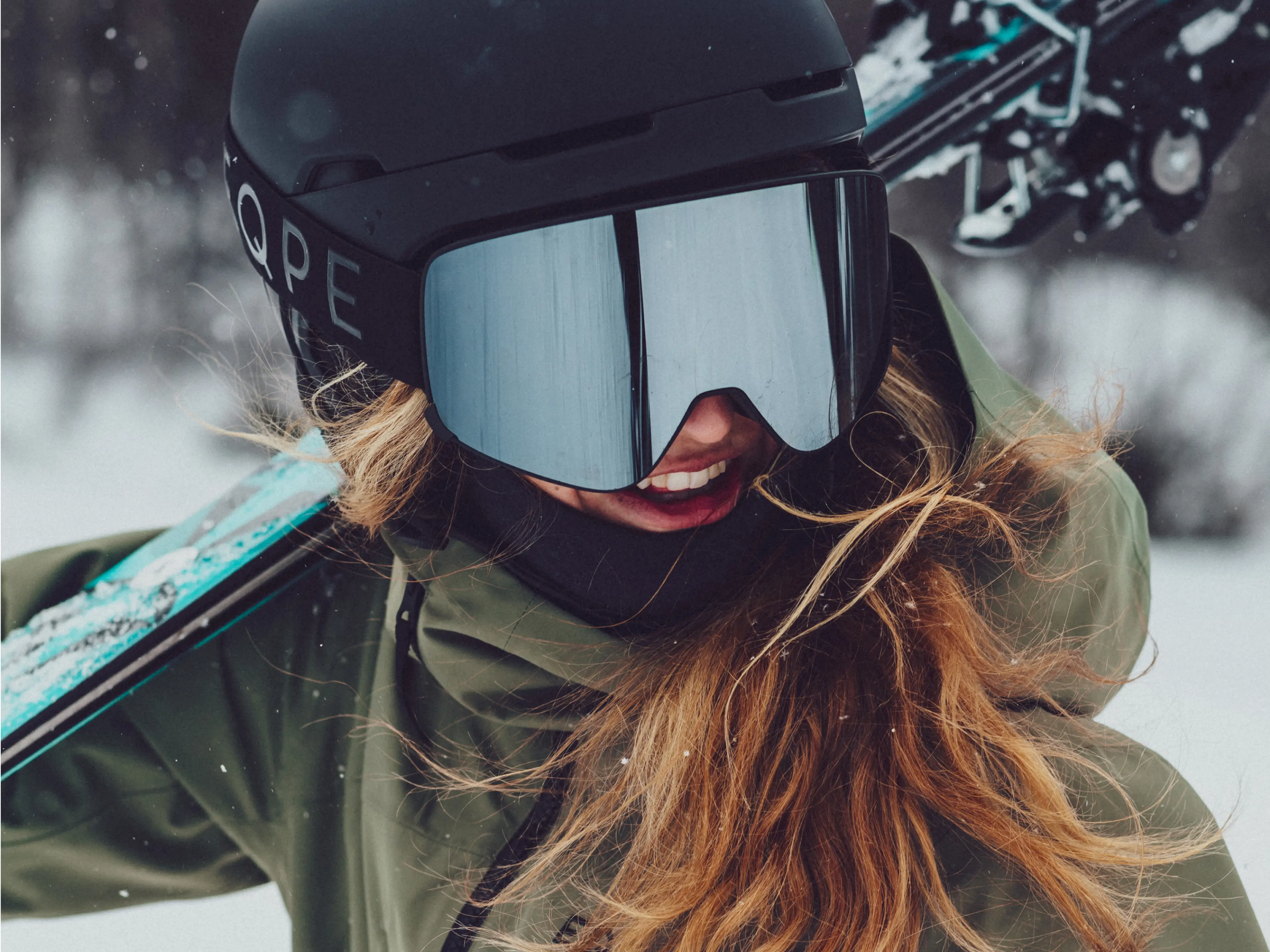 Person in a green jacket and helmet smiling, carrying skis on their shoulder in a snowy setting.