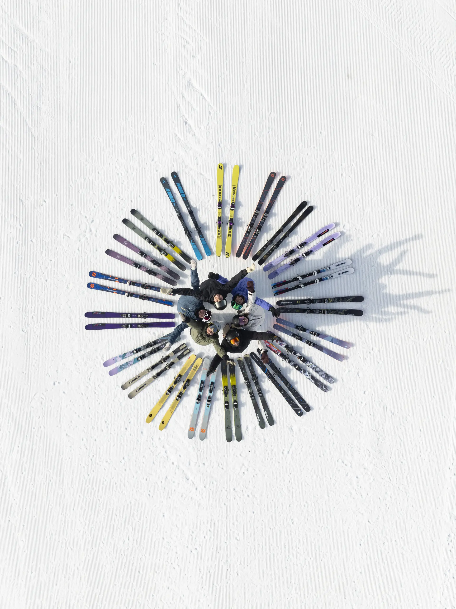 Aerial view of people standing in a circle on snow, heads together, surrounded by skis radiating outward like a sunburst pattern.