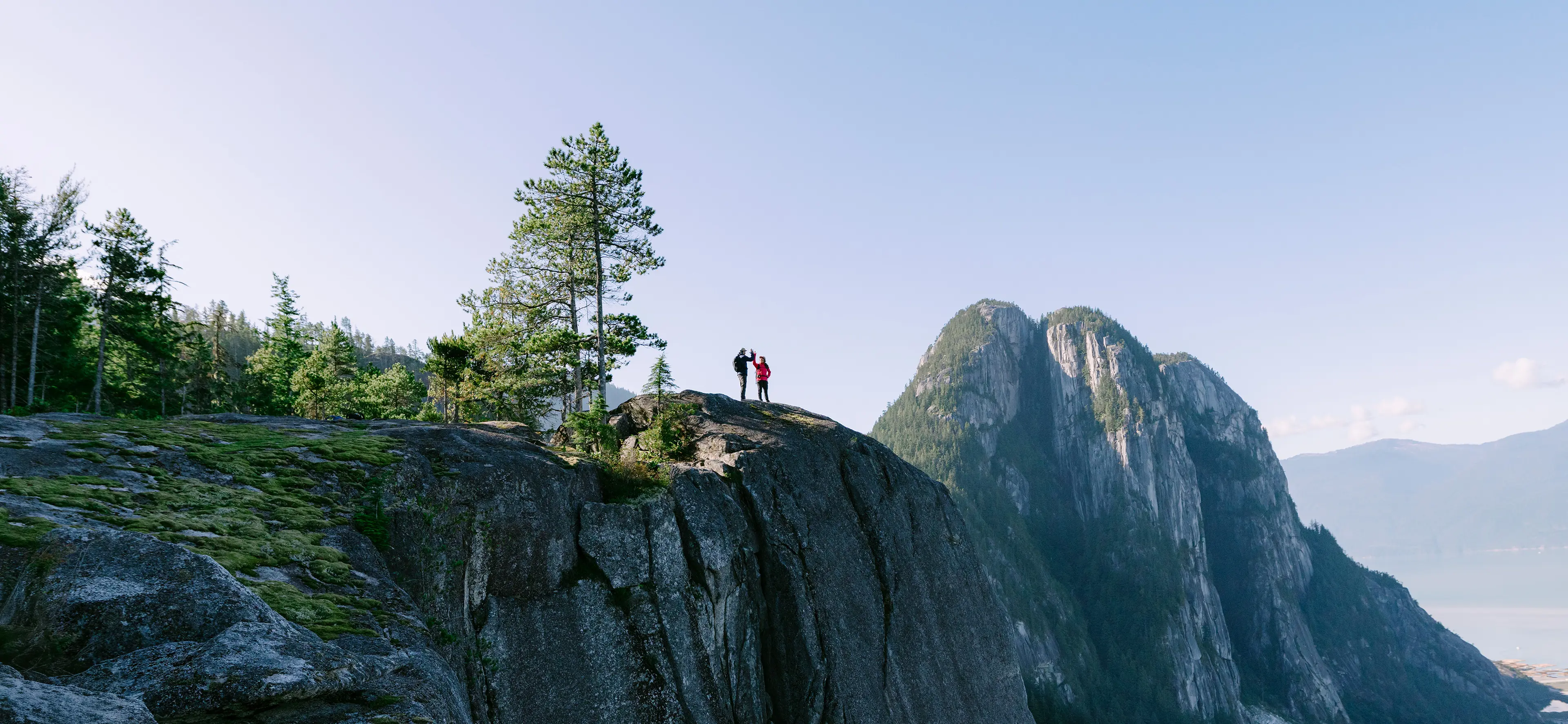 Two hikers wave atop a rocky cliff, surrounded by trees and distant mountains under a clear blue sky.