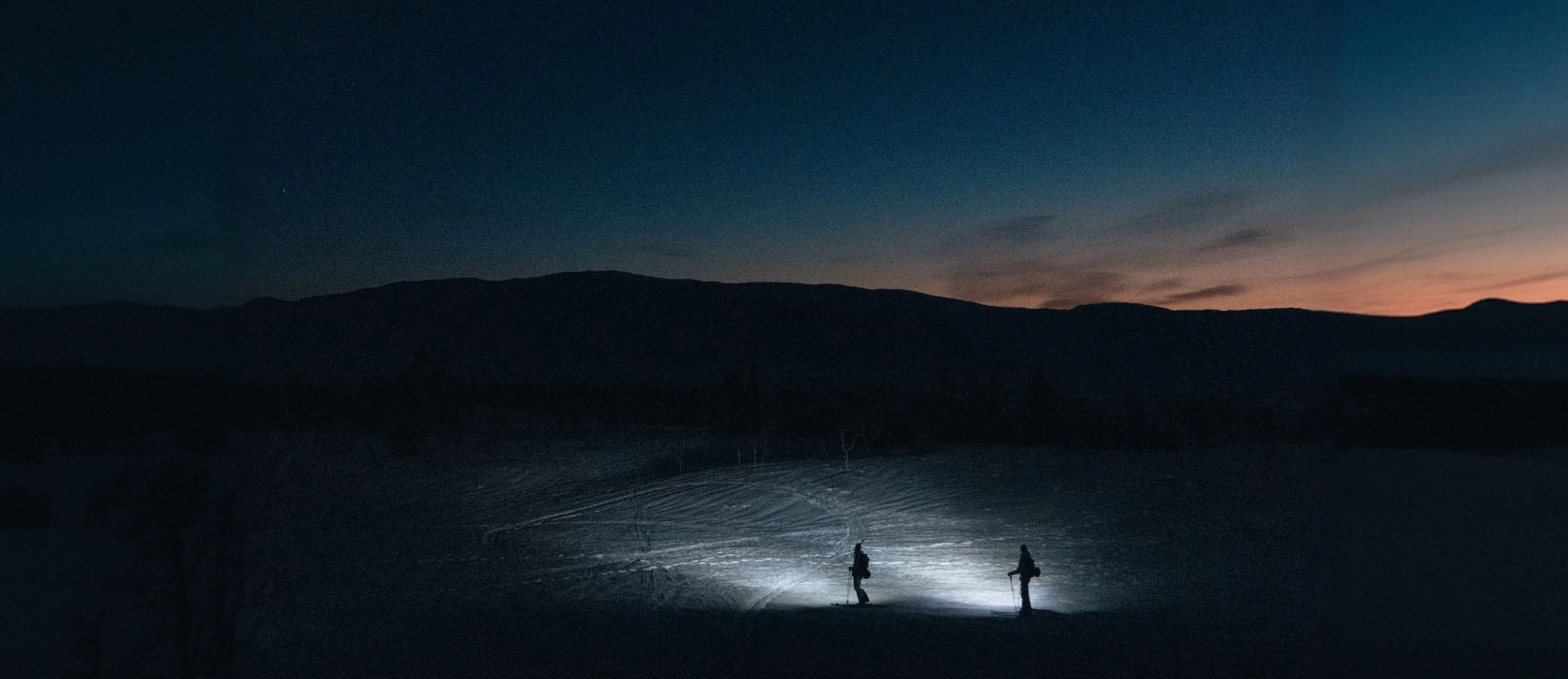 Two people skiing at night with headlamps, surrounded by snow-covered landscape, under a dark blue and orange sky.