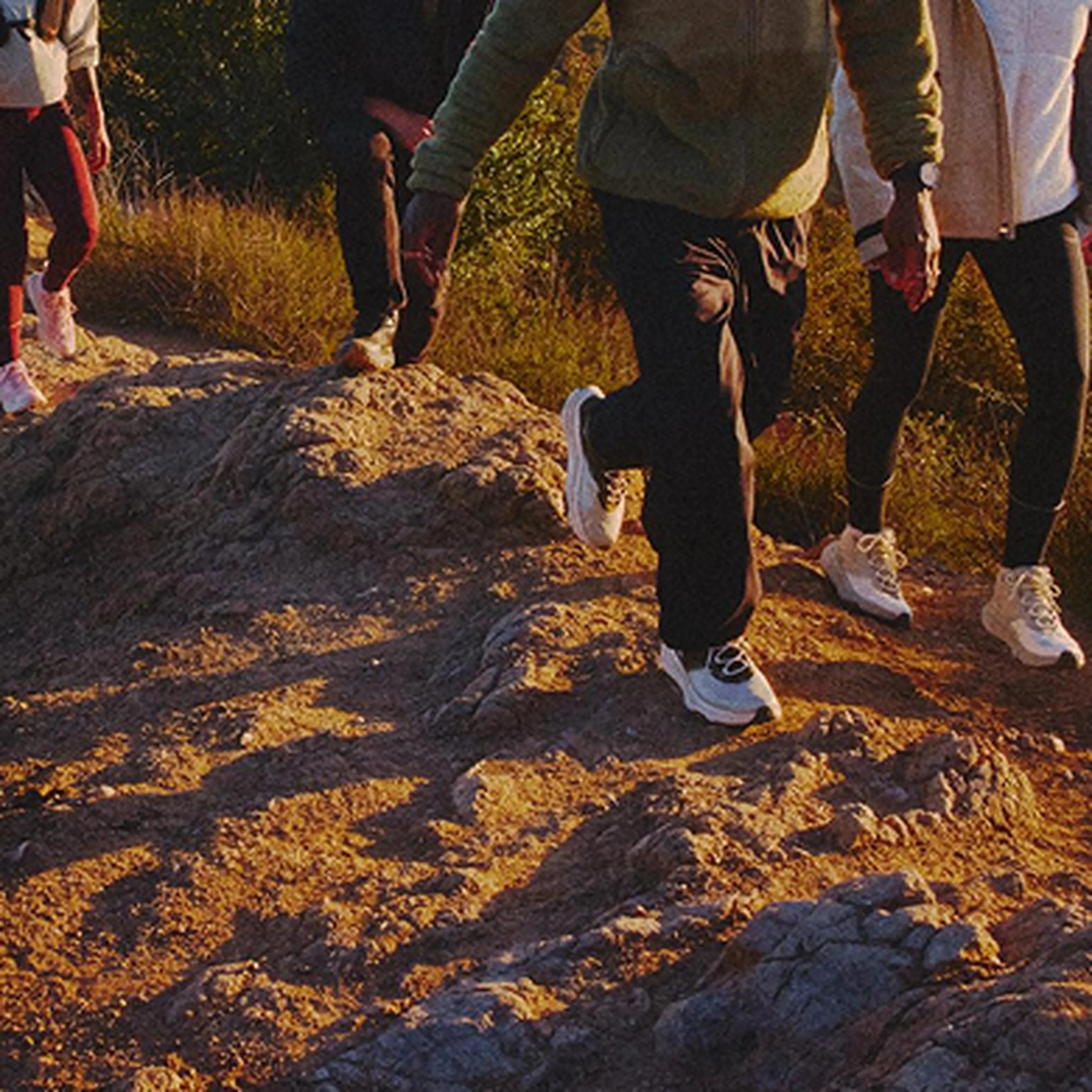 People hiking on a rocky dirt path, wearing casual outdoor clothes and sneakers, with sunlight casting shadows.