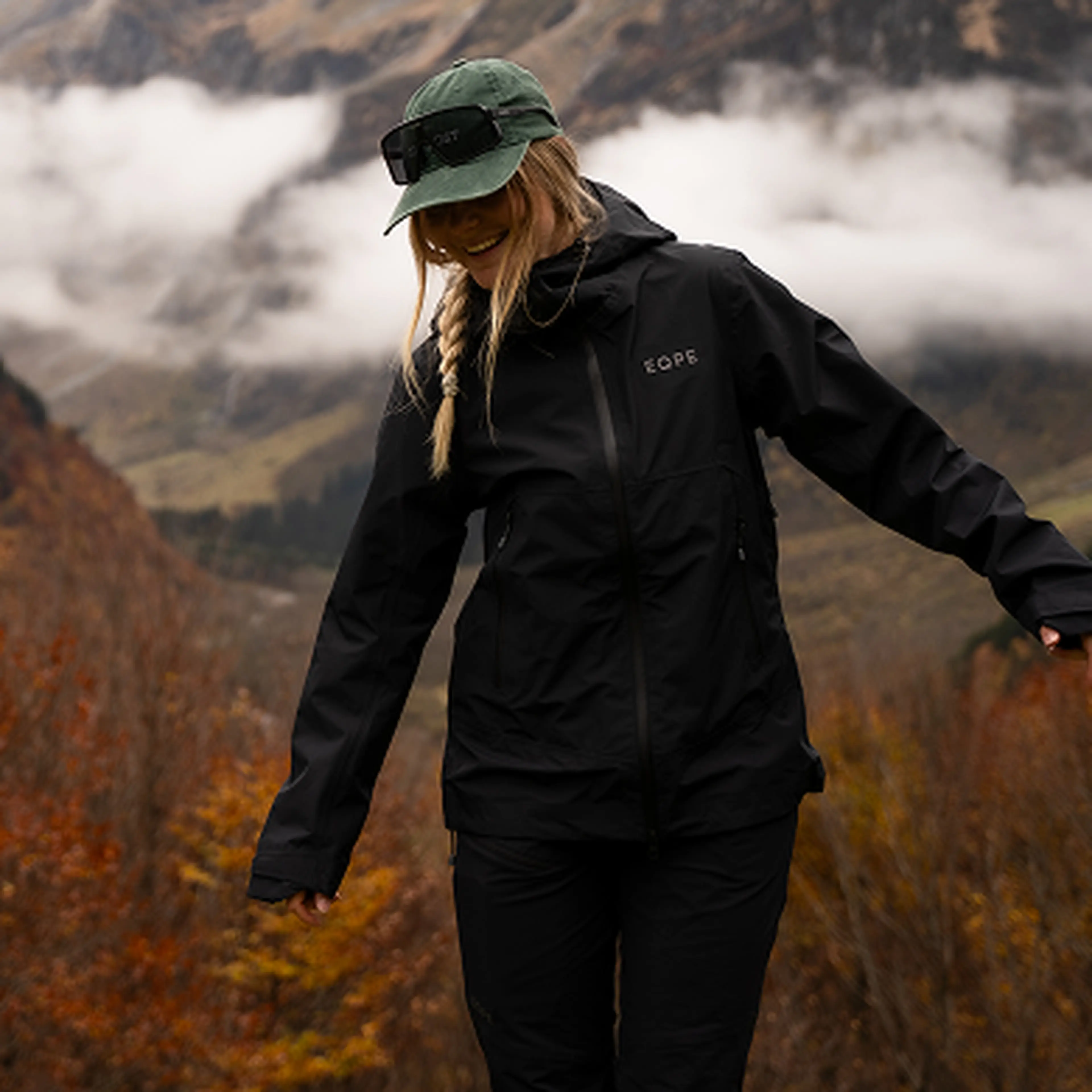 Woman in a black jacket and green cap smiles while standing in a misty mountainous landscape with vibrant autumn foliage.