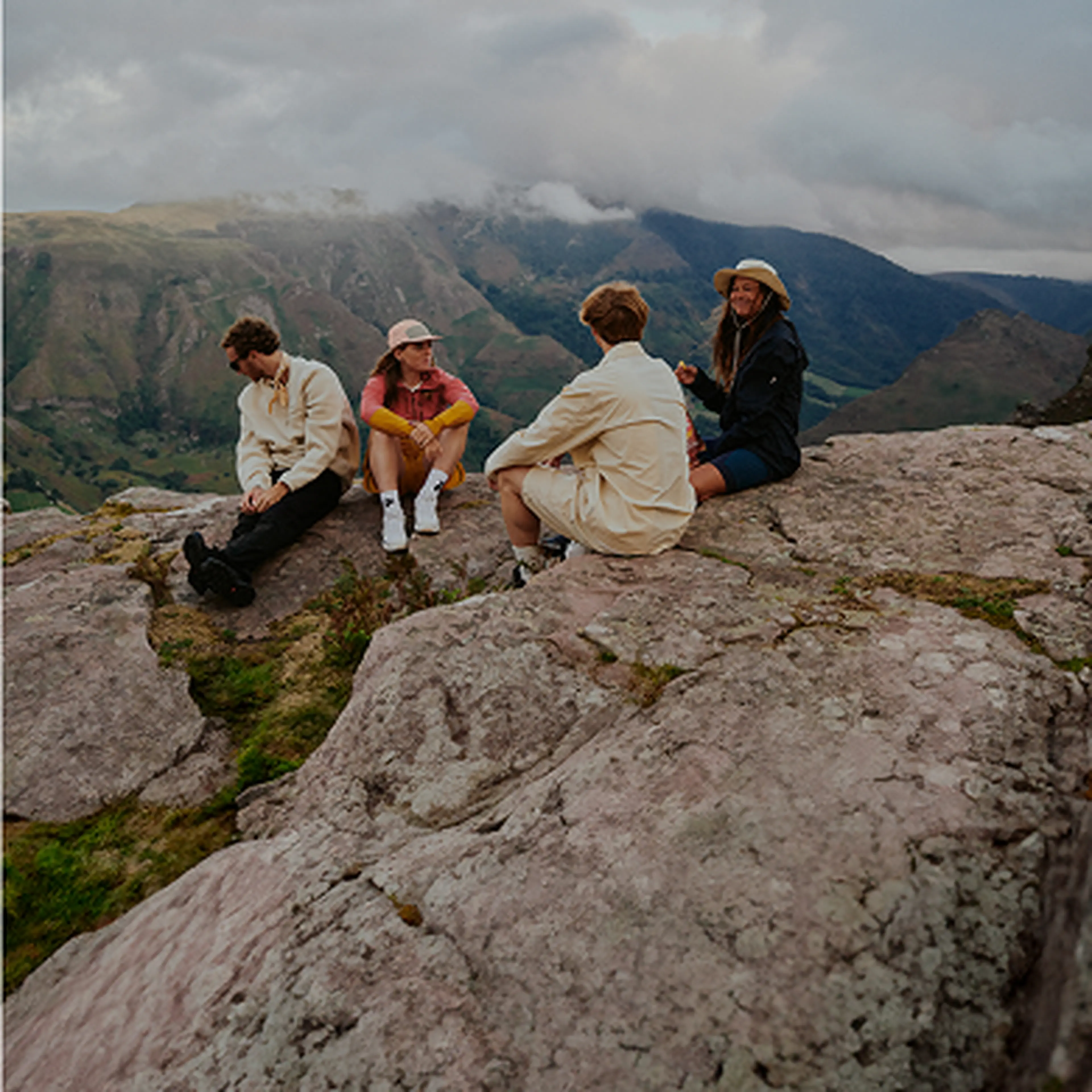 Four people sitting on a rocky mountain ledge, chatting and enjoying a scenic view with cloudy skies and distant hills.