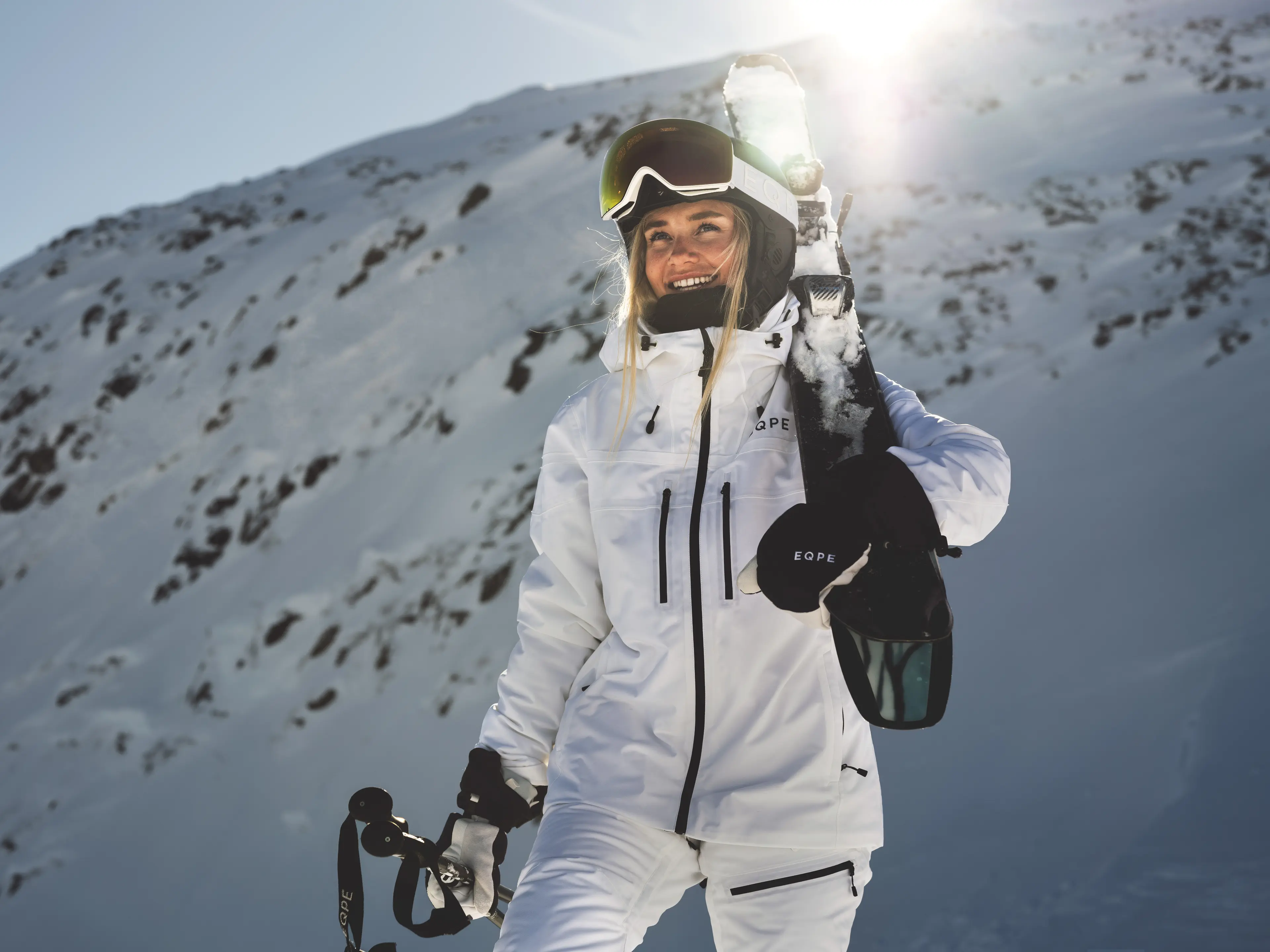 Smiling skier in white gear holds skis and poles, standing on a snowy mountain with sunlight in the background.