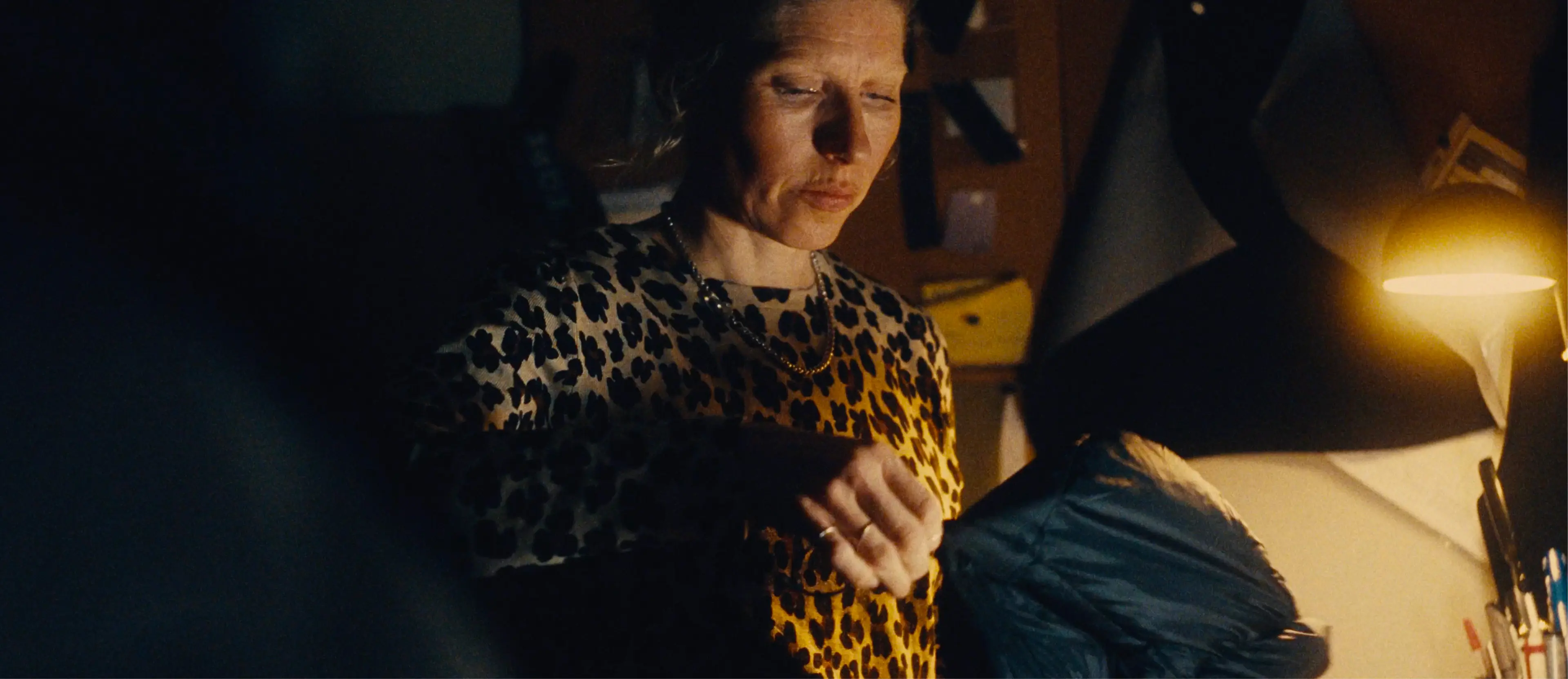 Sara works in a dimly lit room, illuminated by a desk lamp, focusing on a task with concentration.