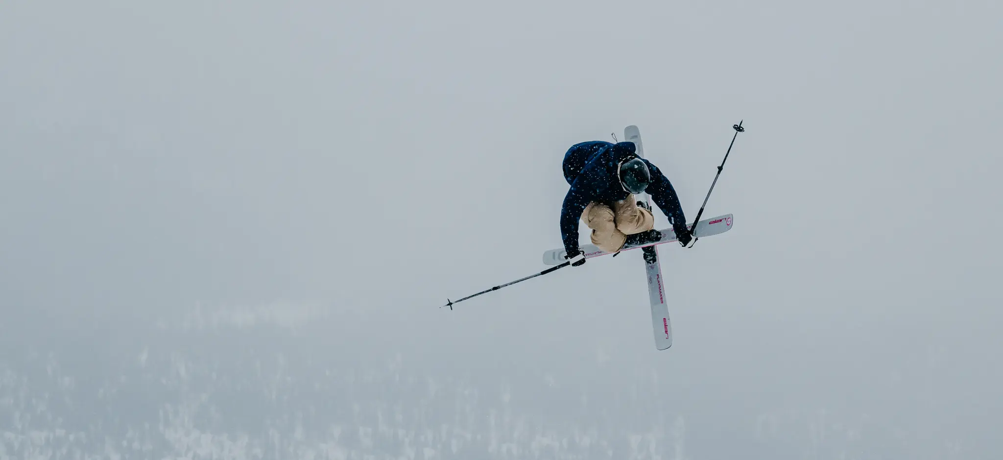 A skier performing an airborne trick with crossed skis against a snowy mountain backdrop, holding ski poles and wearing a helmet.