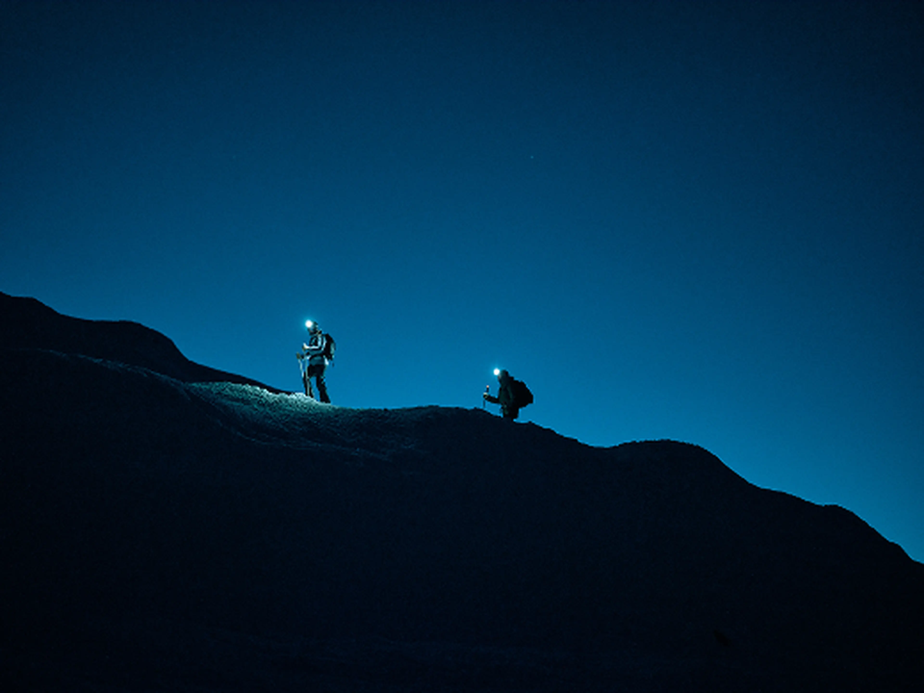 Two hikers with headlamps climb a dark mountain ridge at night, silhouetted against a deep blue sky.