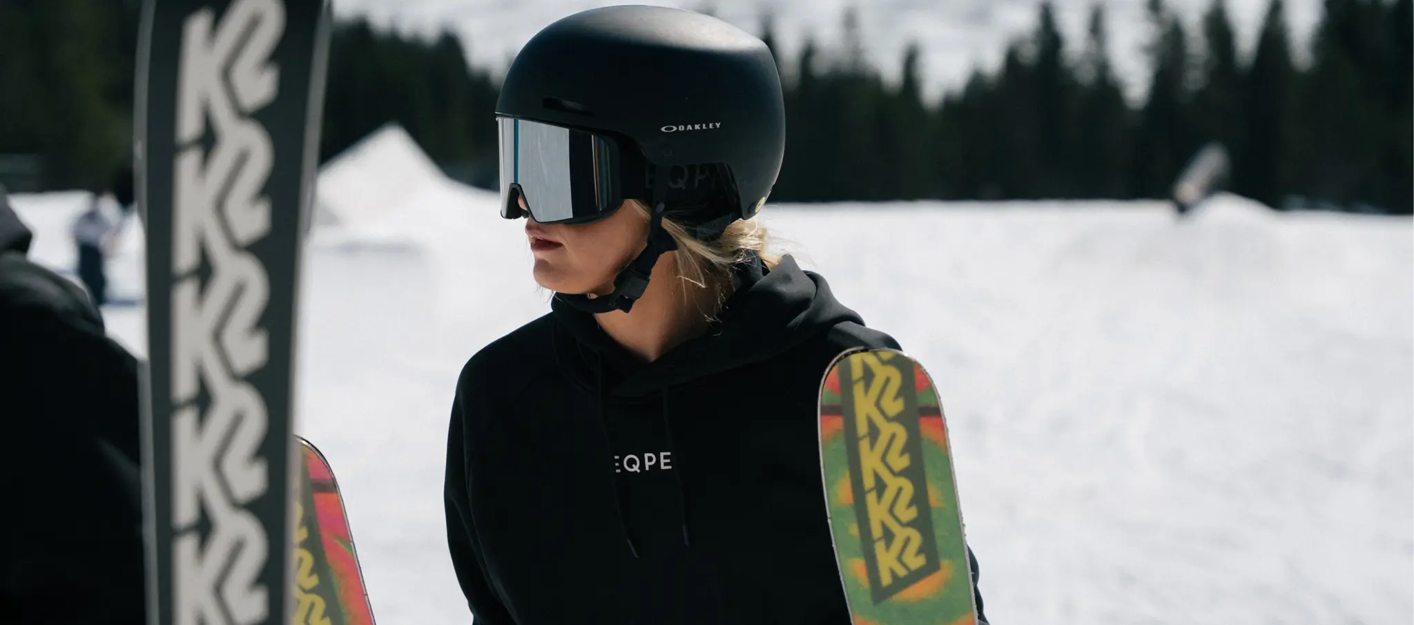 A person in a black helmet and goggles stands on a snowy mountain holding colorful skis, with trees in the background.