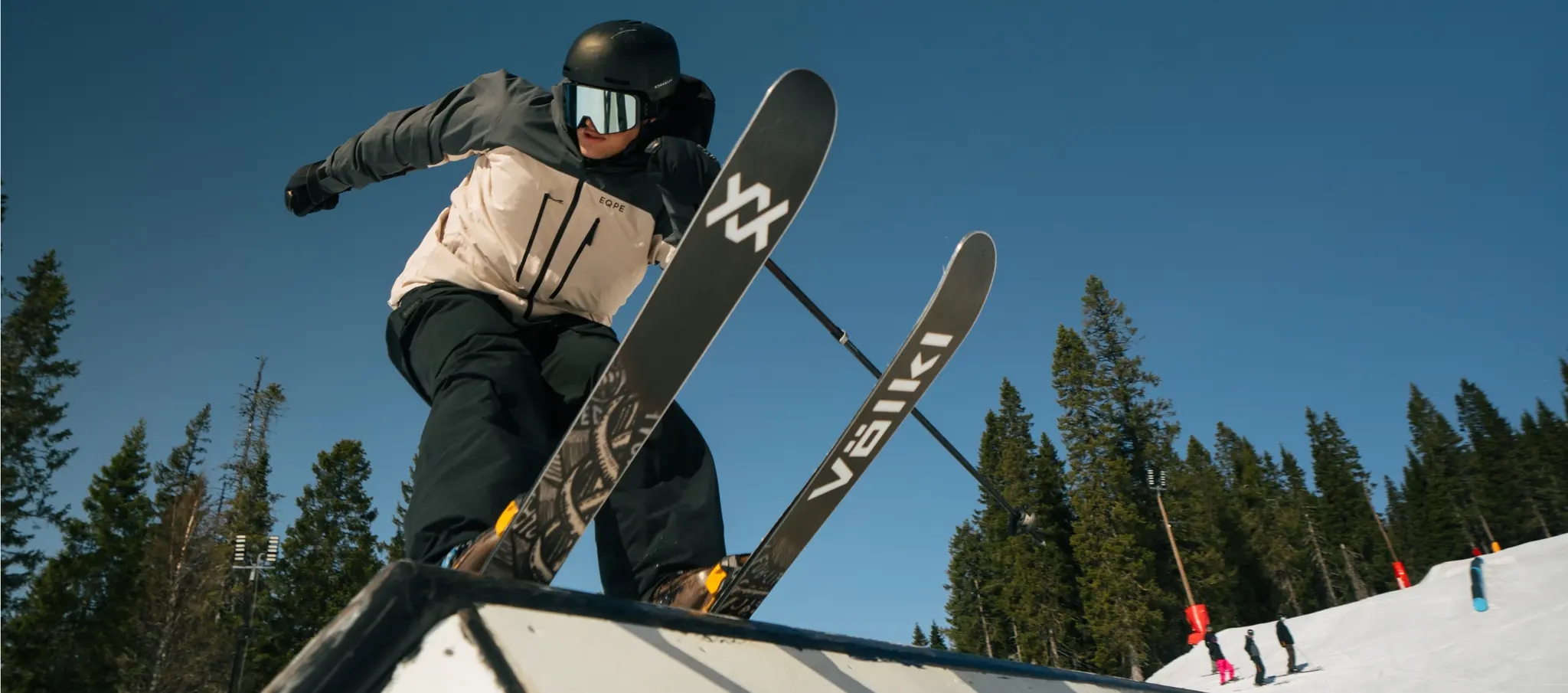 A skier in a black helmet performs a trick on a snowy ramp with a bright blue sky and trees in the background.