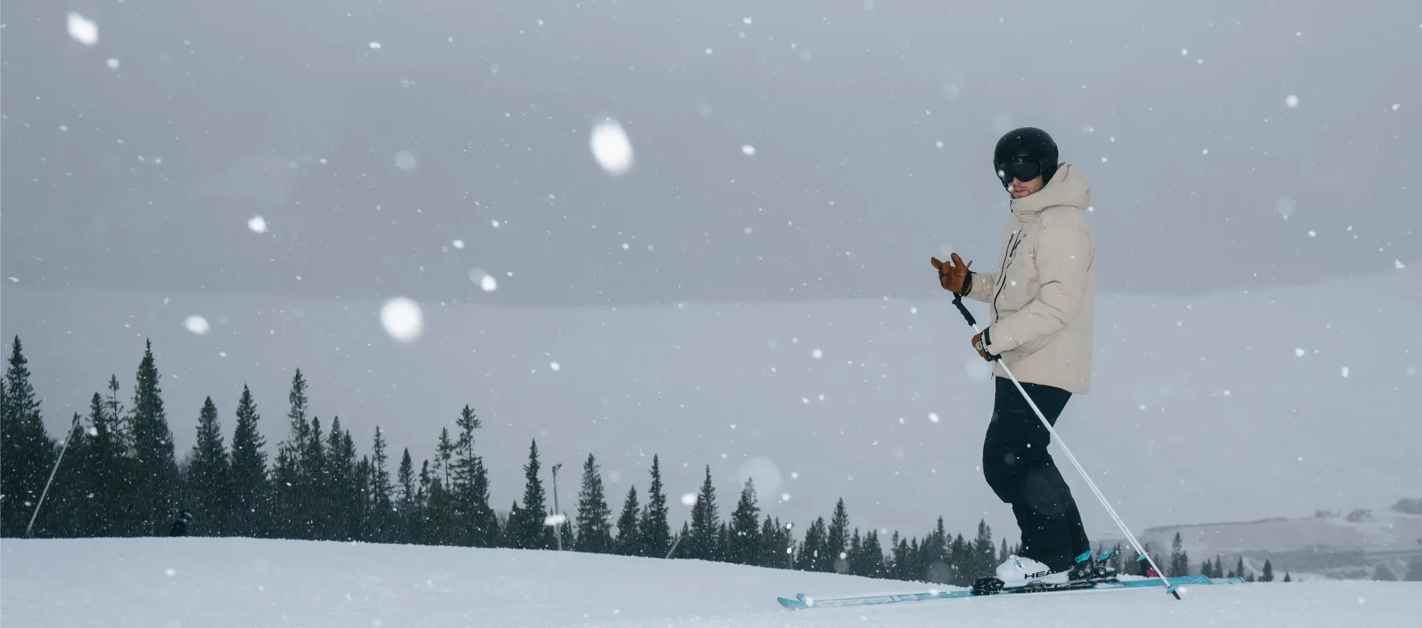 A skier in a beige jacket and helmet stands on a snowy slope with pine trees in the background, while snow falls gently around.