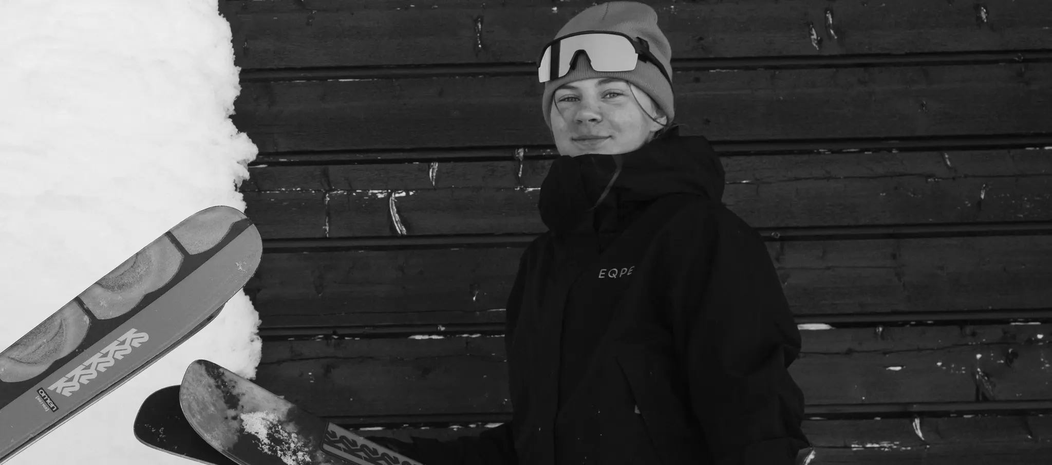 Person wearing a beanie and goggles stands next to skis and a wooden wall, with snow in the background.