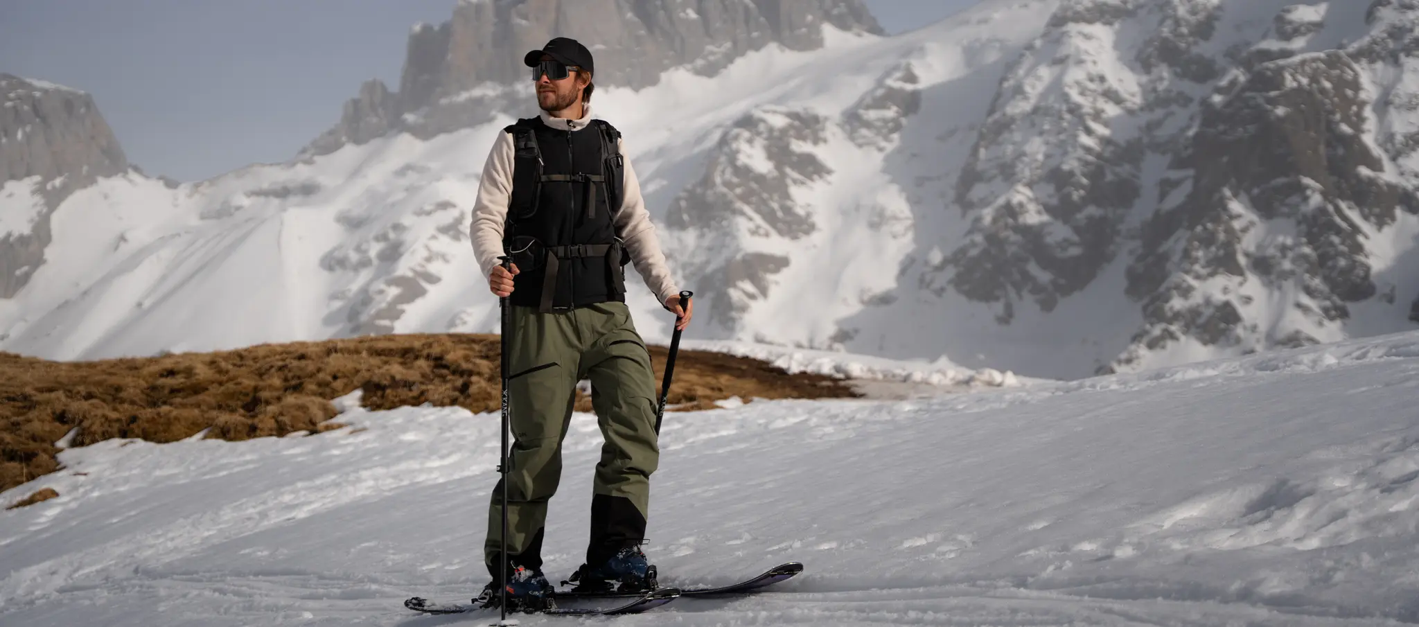 A person in a black vest and green pants stands on skis, holding poles, with snow-covered mountains in the background.
