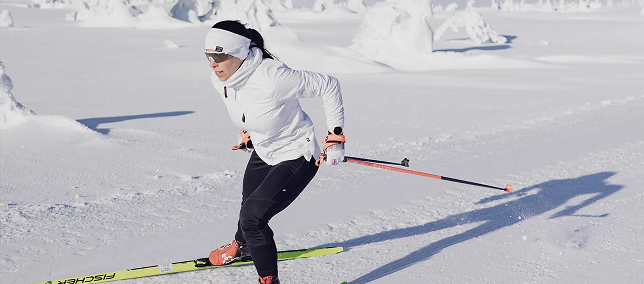 A person wearing a white jacket and headband is cross-country skiing on a snowy landscape under clear skies.