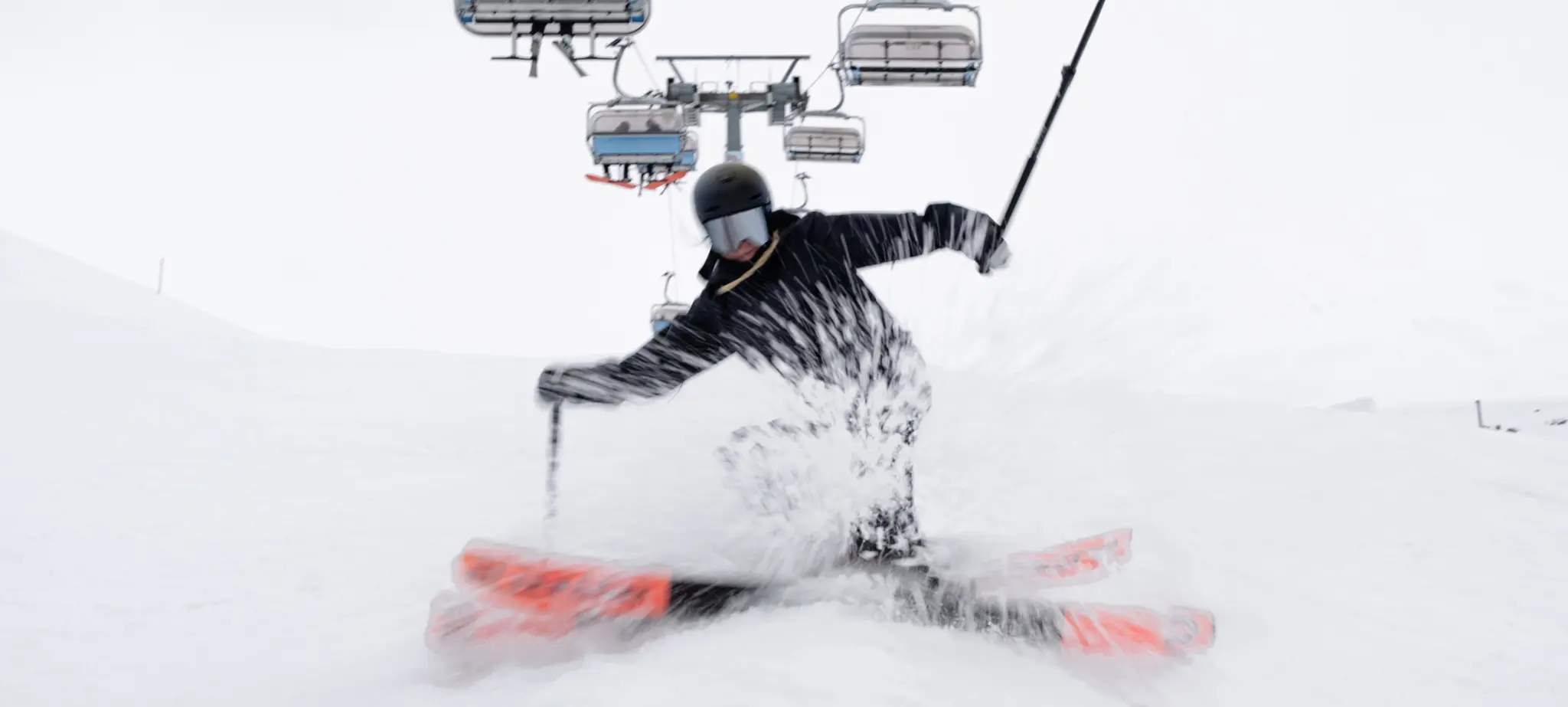 A skier in a black outfit glides down a snowy slope, creating a spray of snow, with ski lifts visible in the background.