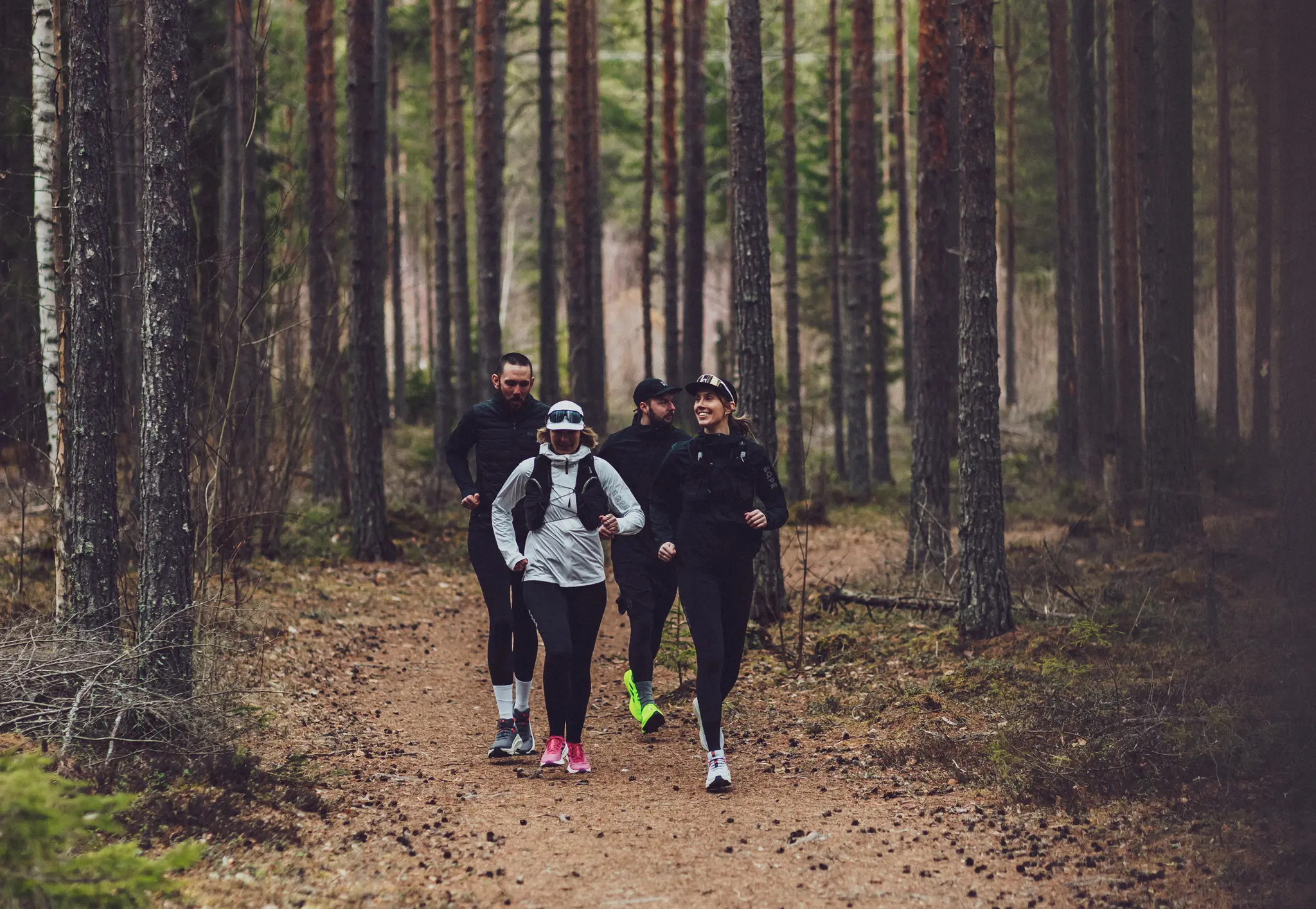 Four people jogging on a forest trail, surrounded by tall trees, wearing athletic gear and sunglasses.