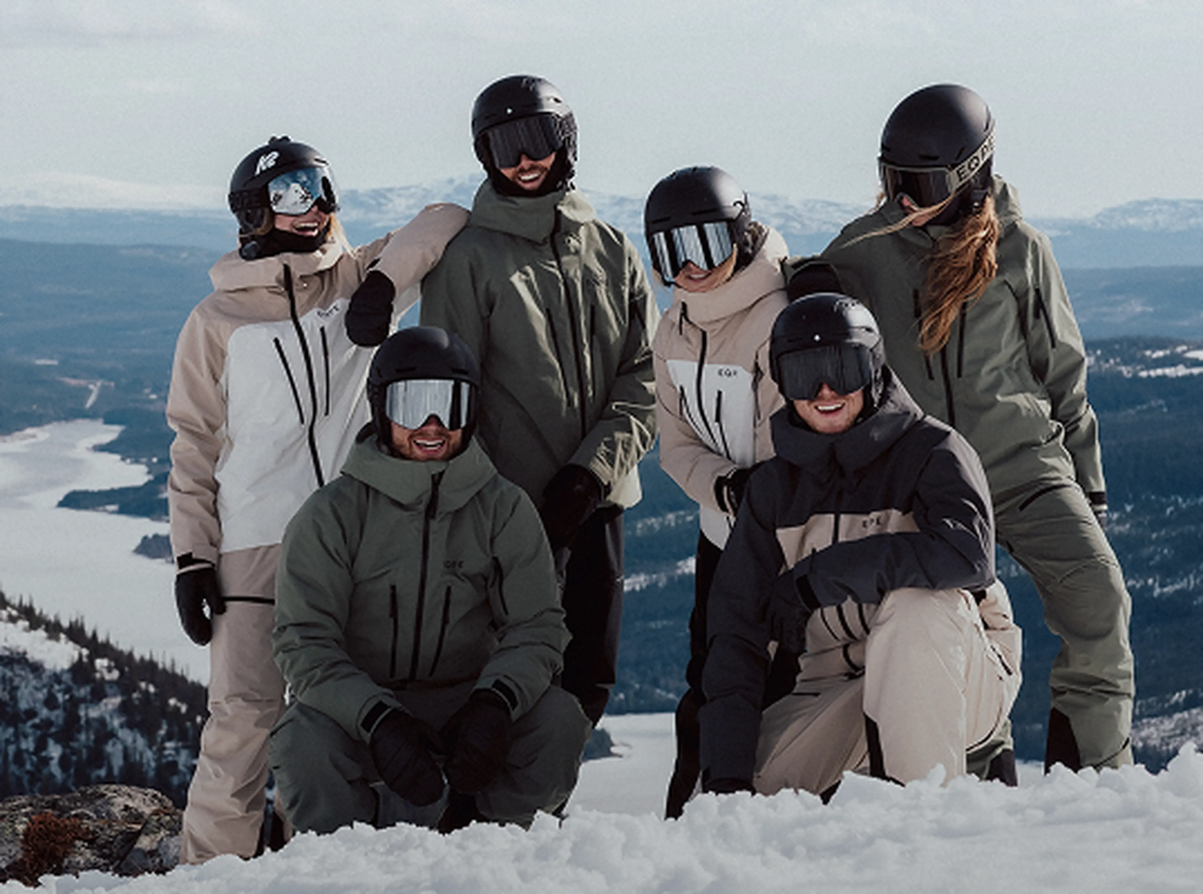A group of six people, wearing helmets and winter gear, pose together on a snowy mountain with a scenic view of distant snow-covered hills.