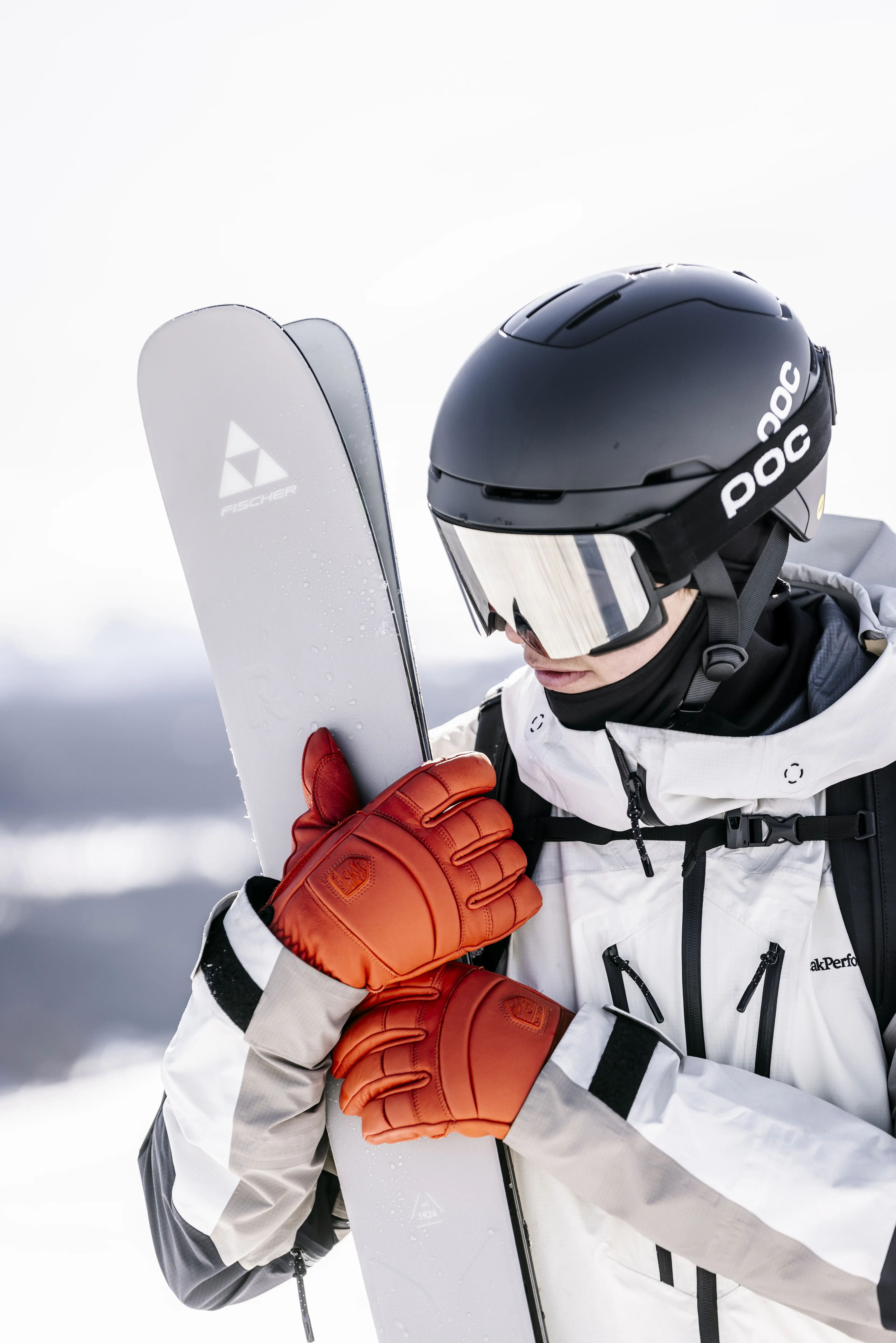 A skier in a white jacket and orange gloves holds a pair of skis in a snowy landscape.