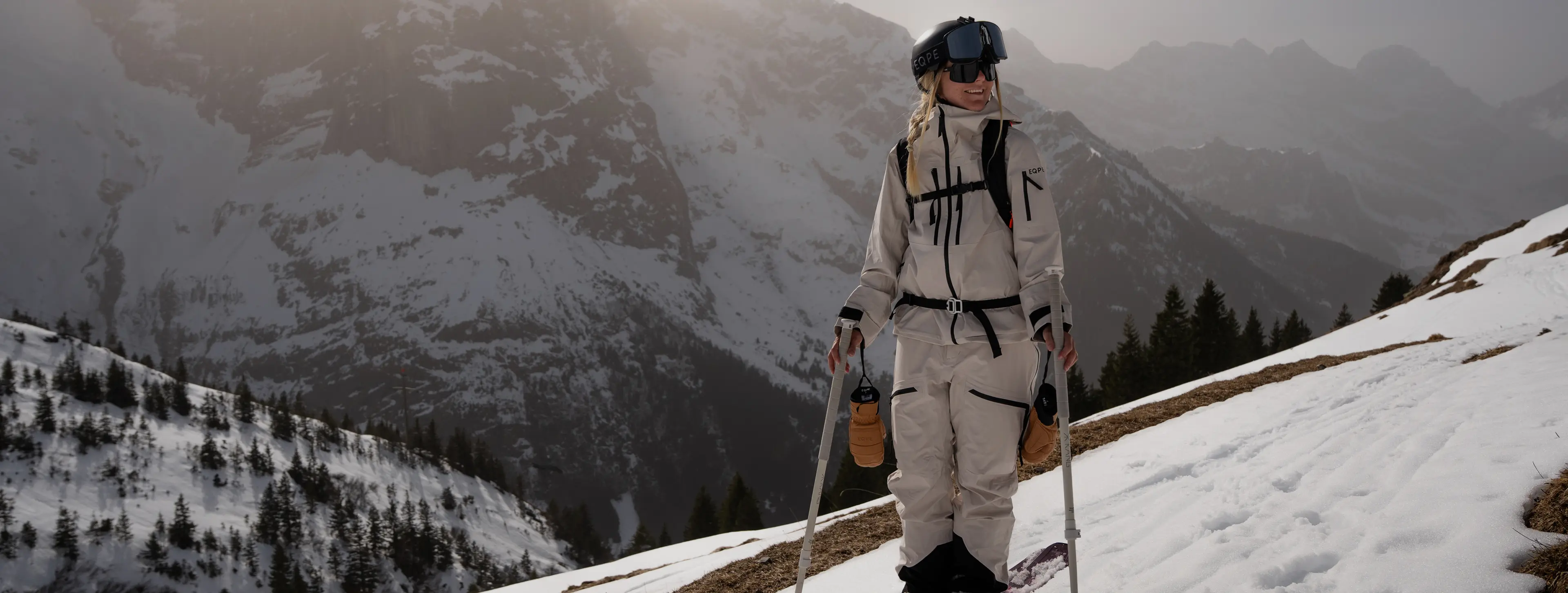 Person in snow gear with ski poles stands on a snowy mountain slope, surrounded by misty, mountainous landscape.