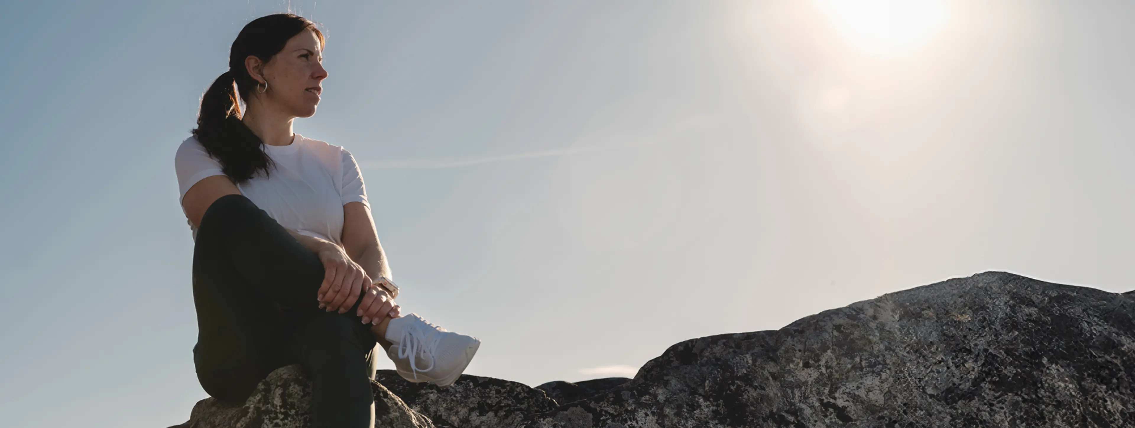A person in a white shirt and ponytail sits on a rock, looking thoughtfully into the distance under a bright sun in a clear sky.