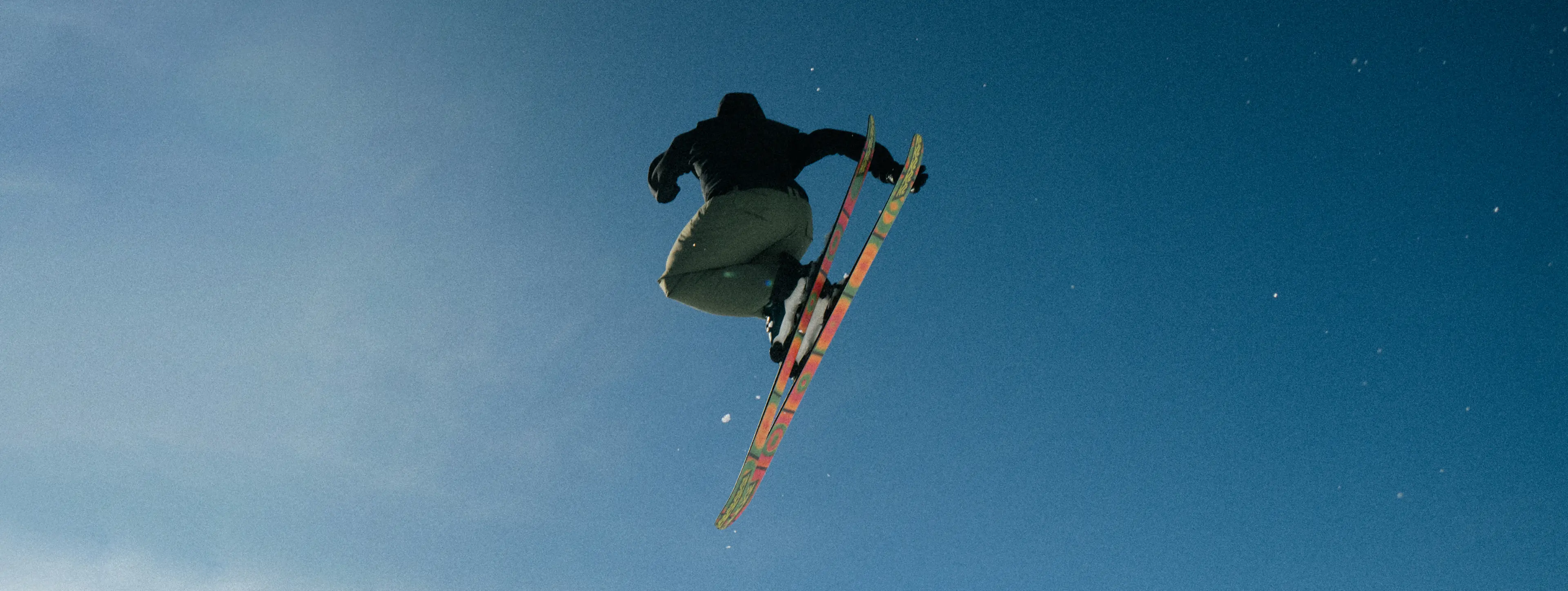 A skier in mid-air performs a jump against a clear blue sky, showcasing colorful skis and dynamic motion.