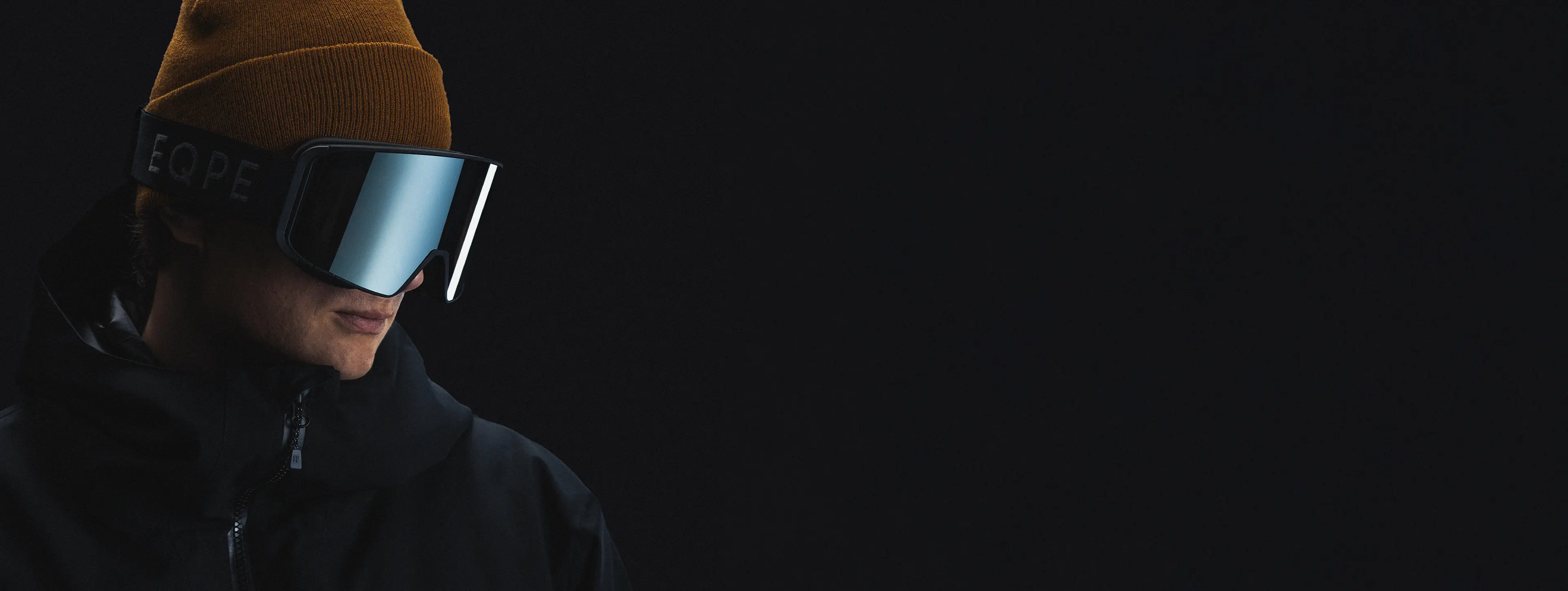 Person wearing a brown beanie, mirrored ski goggles, and a dark jacket against a black background.