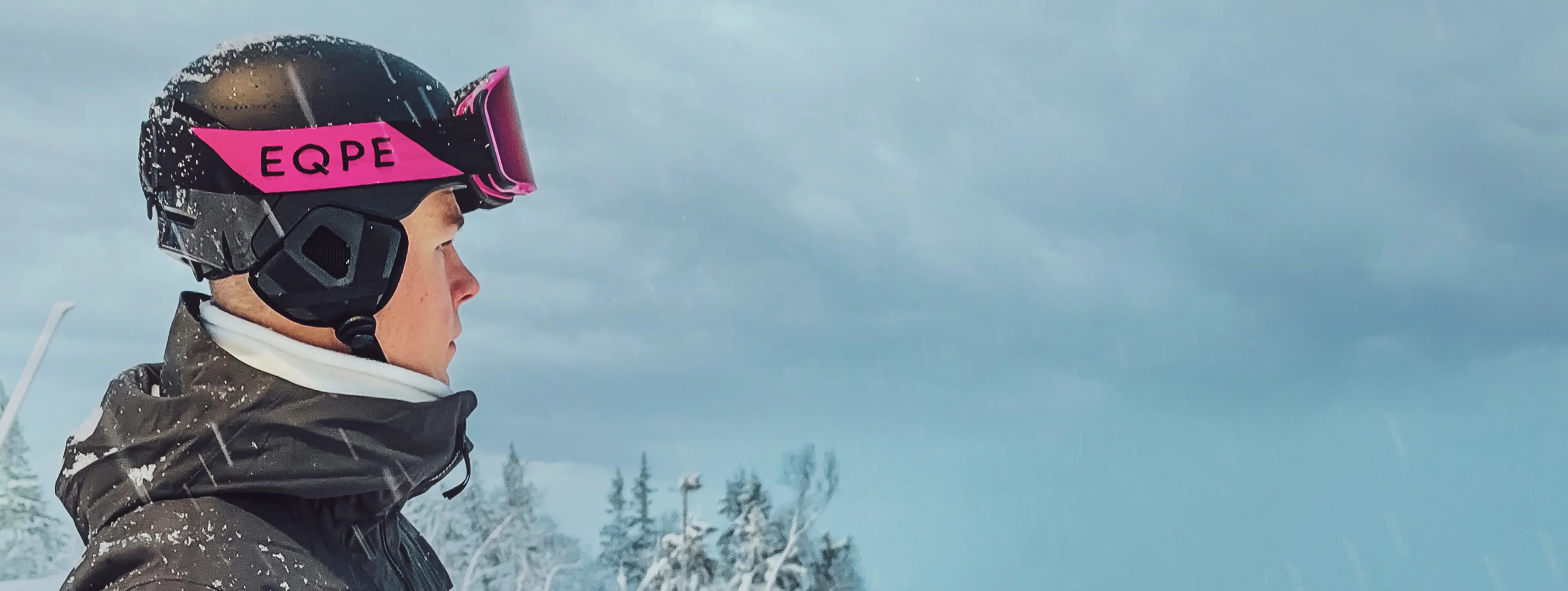 A person wearing a helmet and ski goggles is standing in a snowy landscape, looking towards the distance under a cloudy sky.