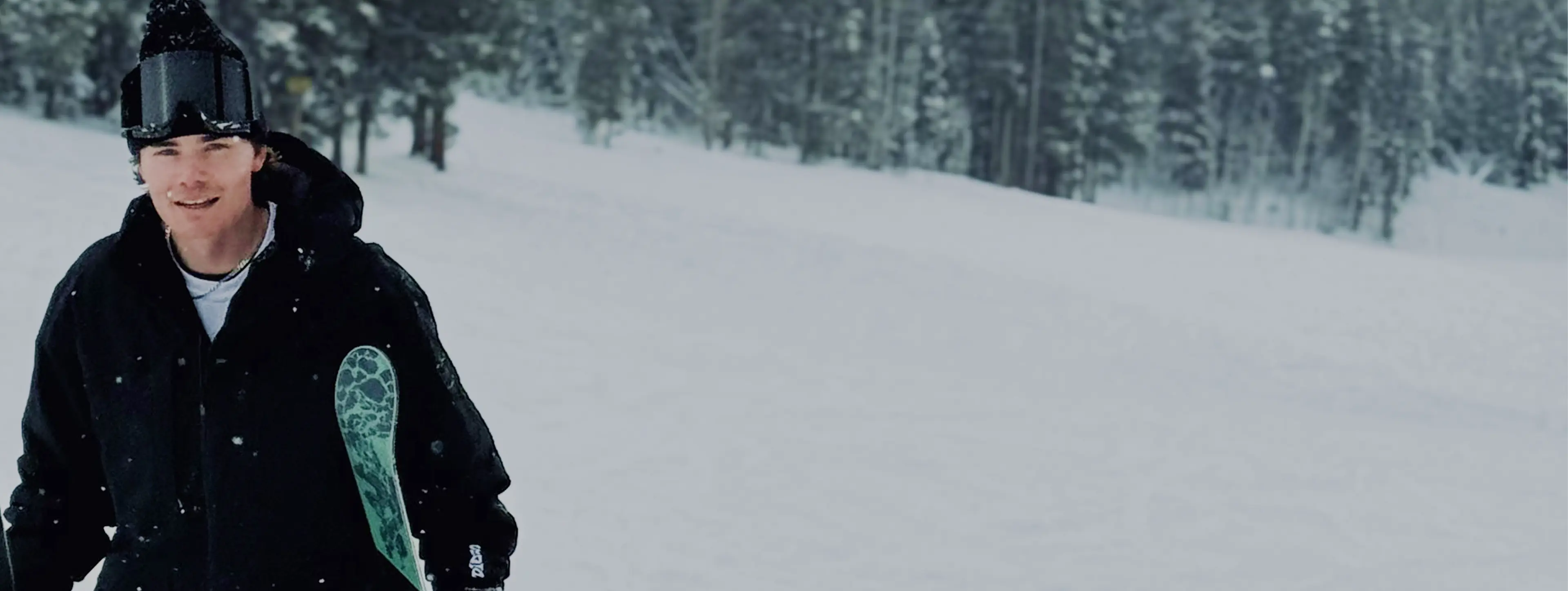 A person in winter gear holding skis, standing on snow-covered terrain with a forest background.