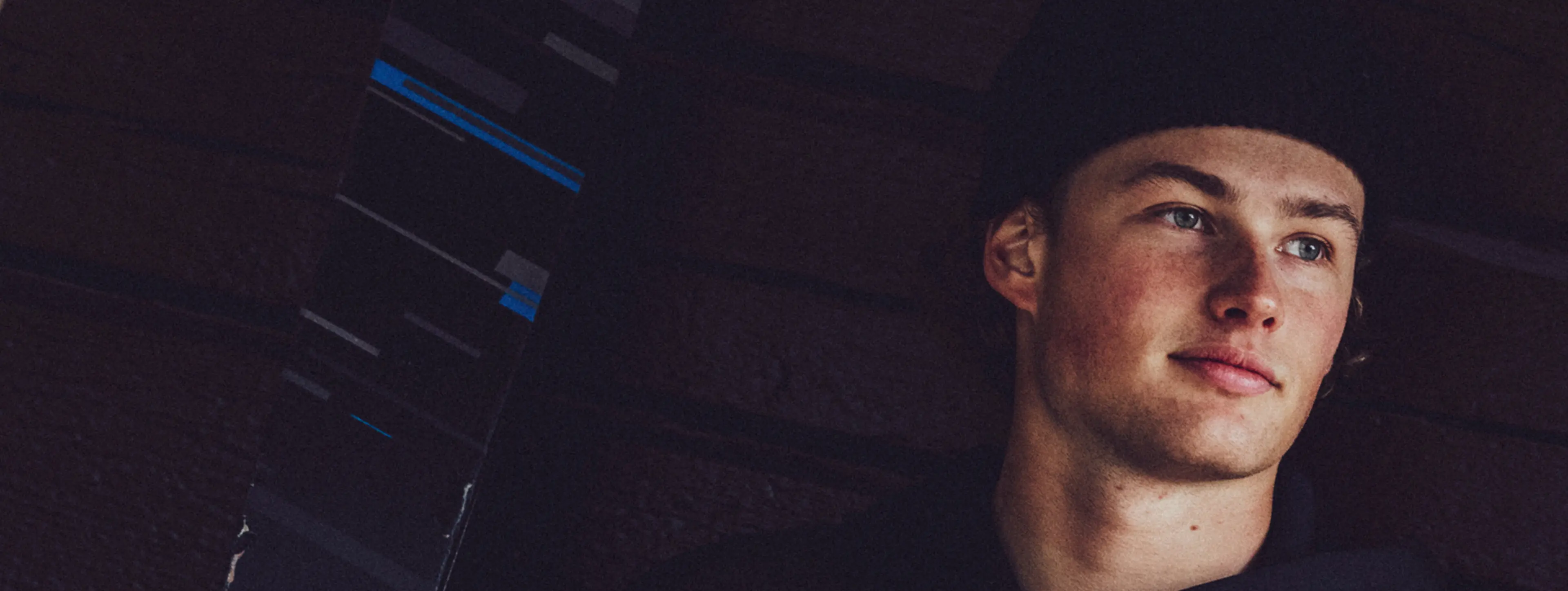 Young man wearing a black beanie, looking off to the side, with a pensive expression against a dark, textured background.